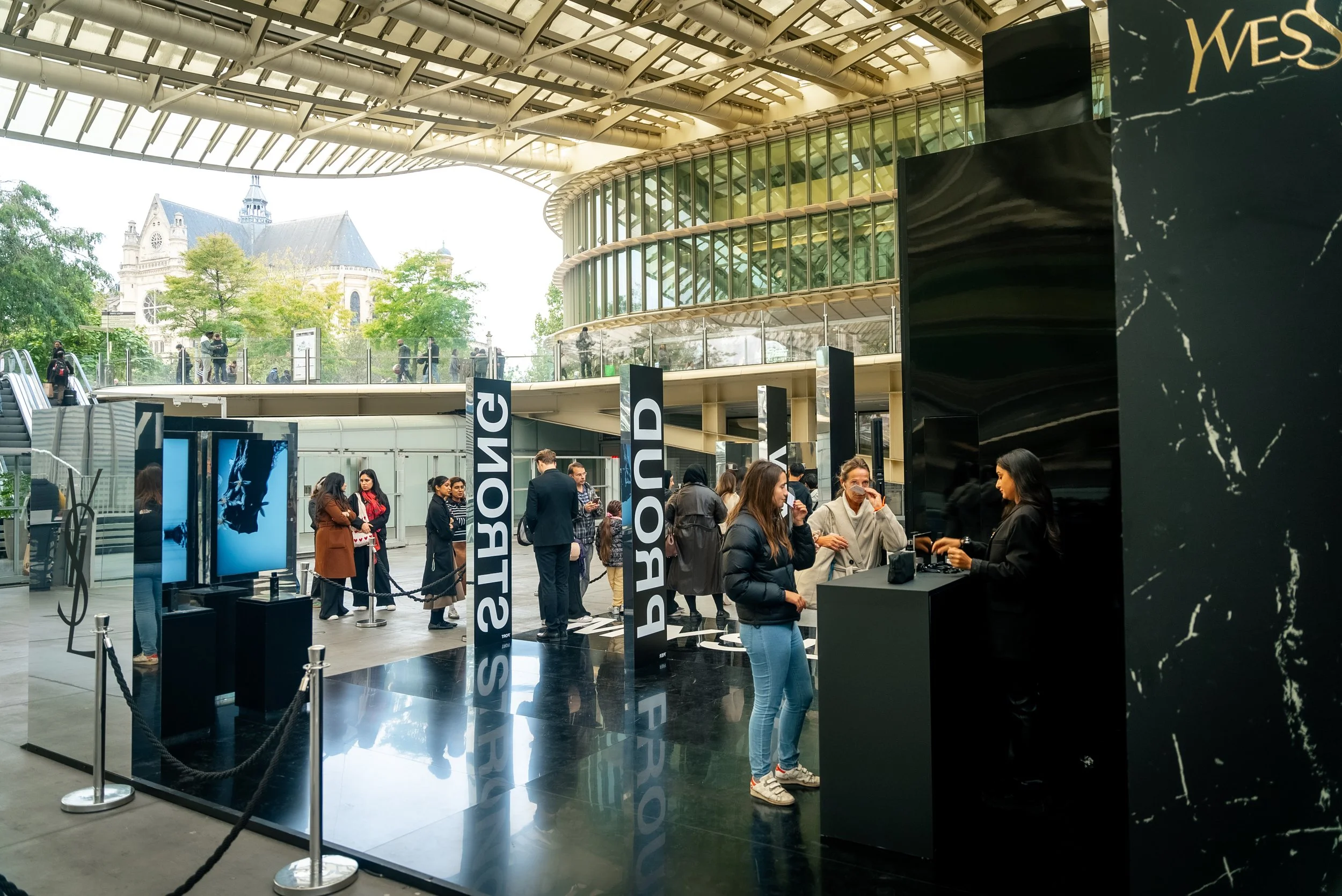 Une file d'attente à l'extérieur d'un bâtiment moderne avec une enseigne affichant 'SOUVENIRS'. Des personnes font la queue, d'autres interagissent avec le personnel du stand, dans un cadre urbain avec des arbres et un bâtiment historique en arrière-
