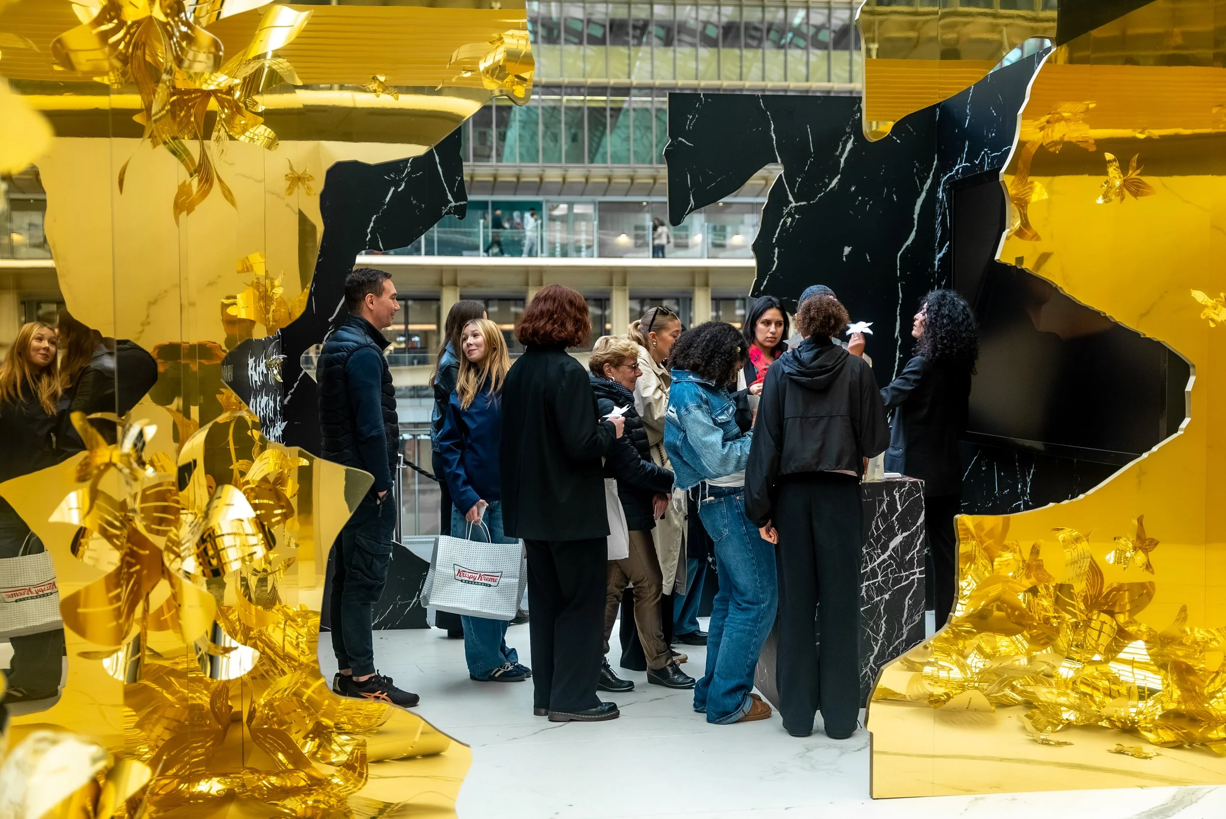 Groupe de personnes faisant la queue à l'intérieur, encadré par des décors en or et noir avec des motifs de marbre, dans un espace moderne avec beaucoup de lumière naturelle.