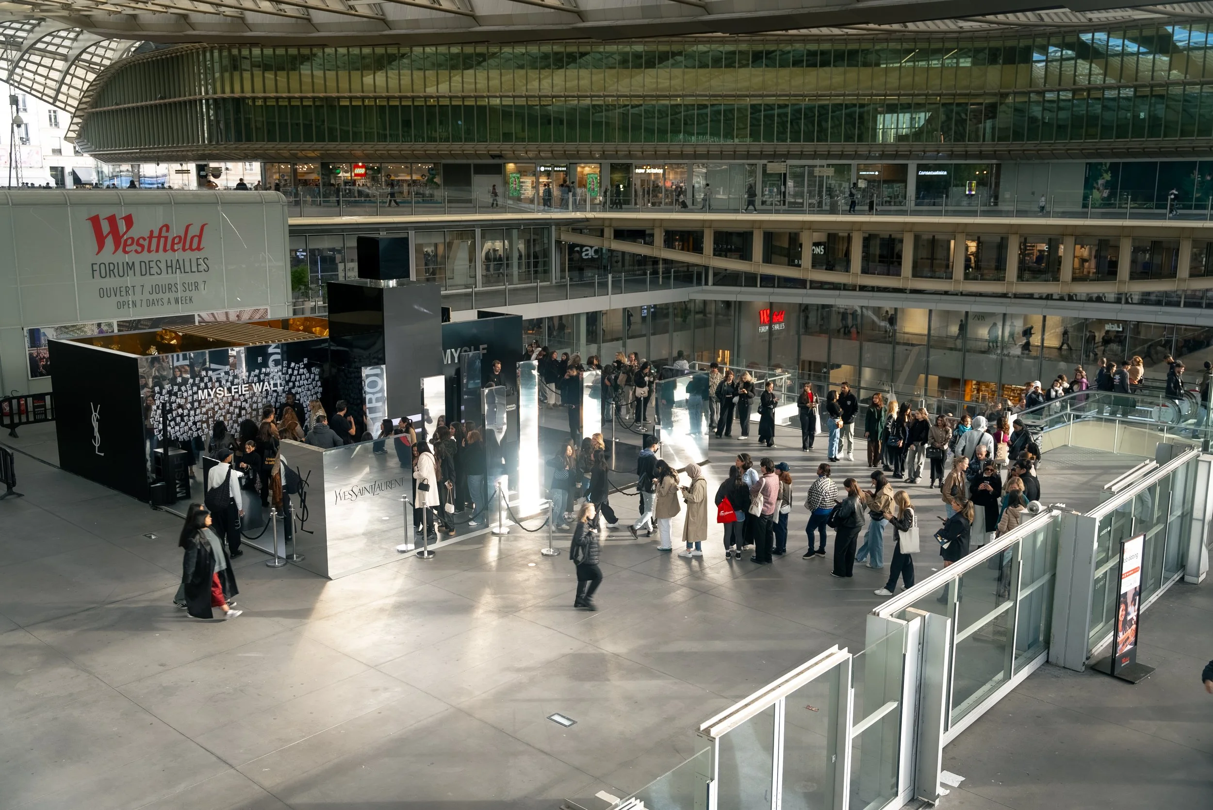 File d'attente de personnes dans un centre commercial ou un bâtiment public, avec un stand de marque Yves Saint Laurent.