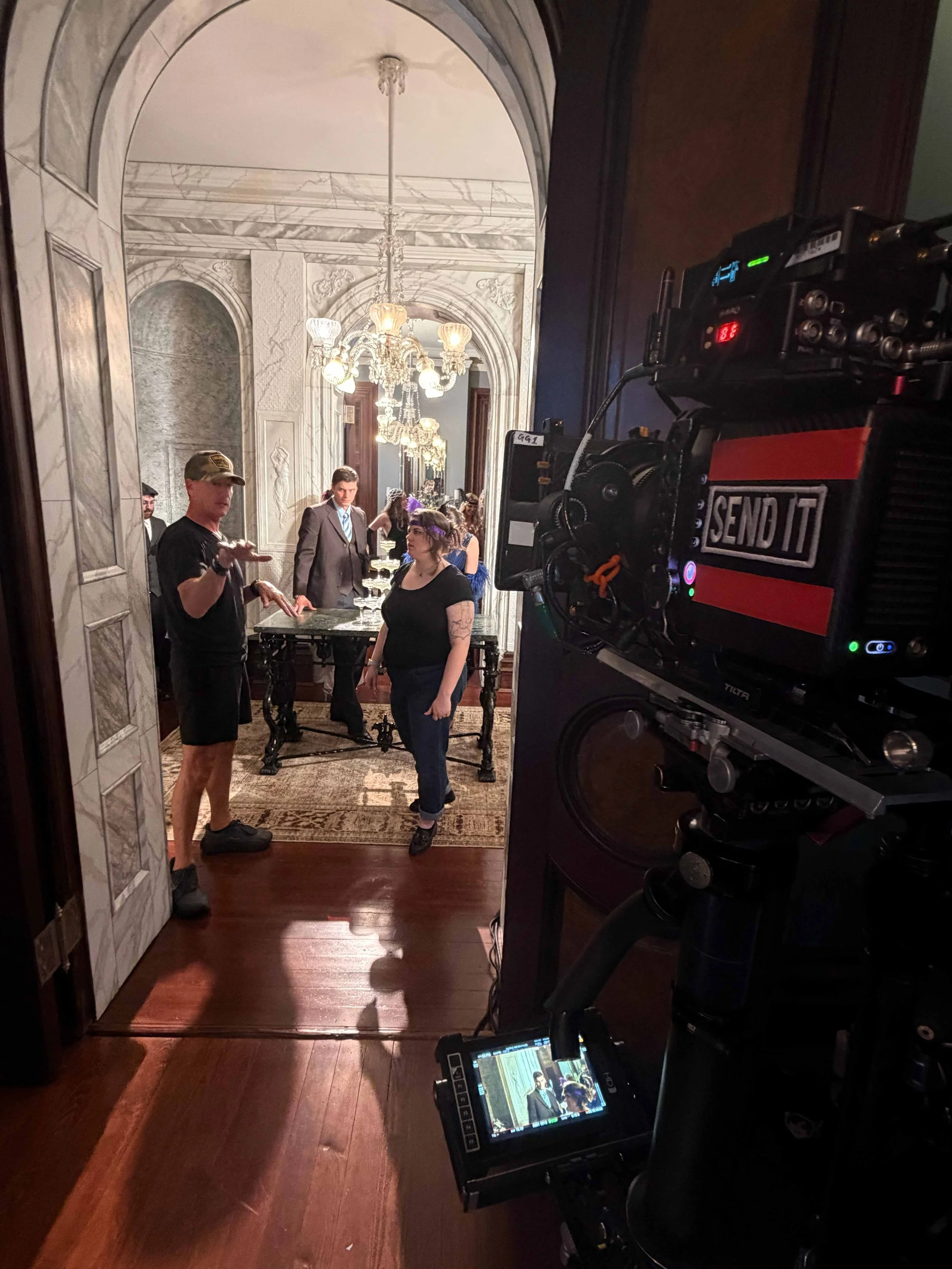 Filming crew setting up a scene in an ornate, marble-finished hallway with chandeliers and intricate wall details, with a camera and monitor in the foreground. Commercial shoot for Hale's Jeweler's, Greenville SC.