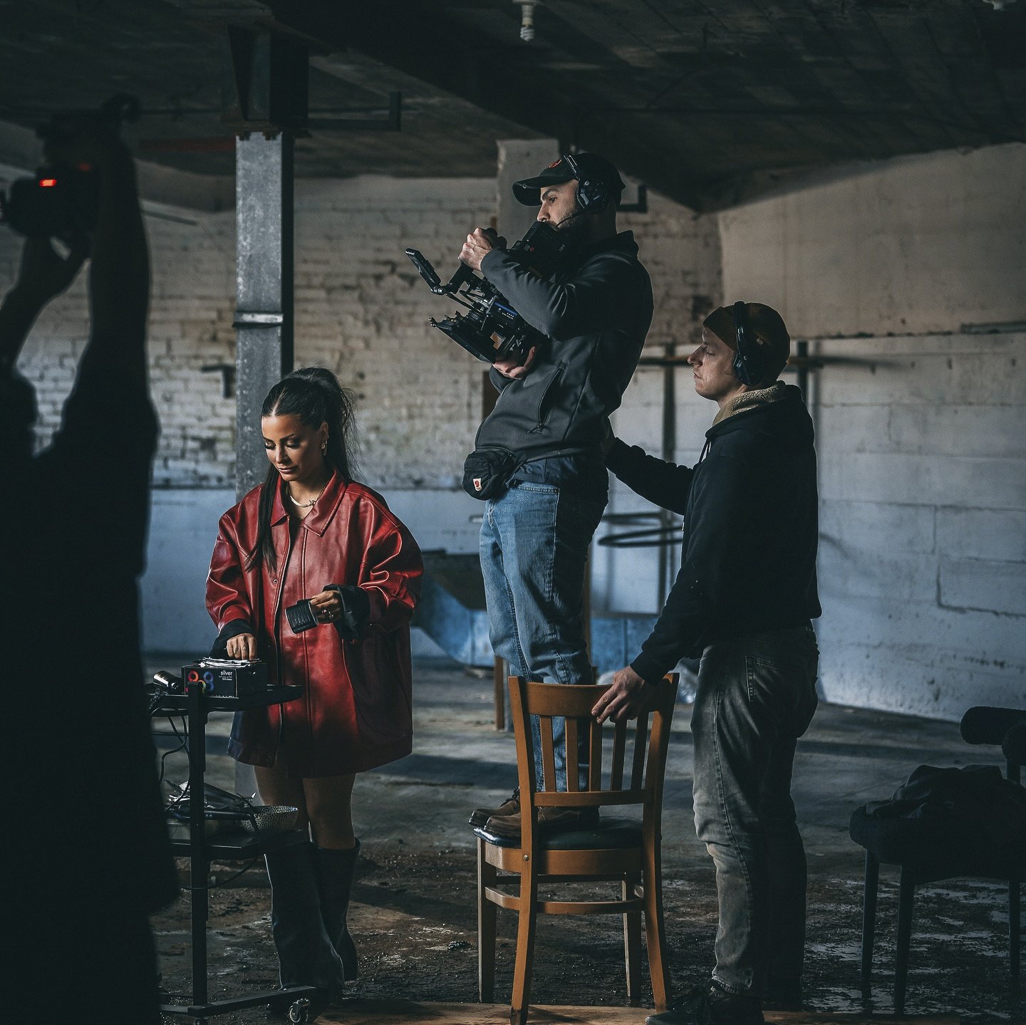 A film crew on set in an industrial space, with a woman in a red leather jacket operating audio equipment, a man with a camera rig standing on a chair, and another man assisting.