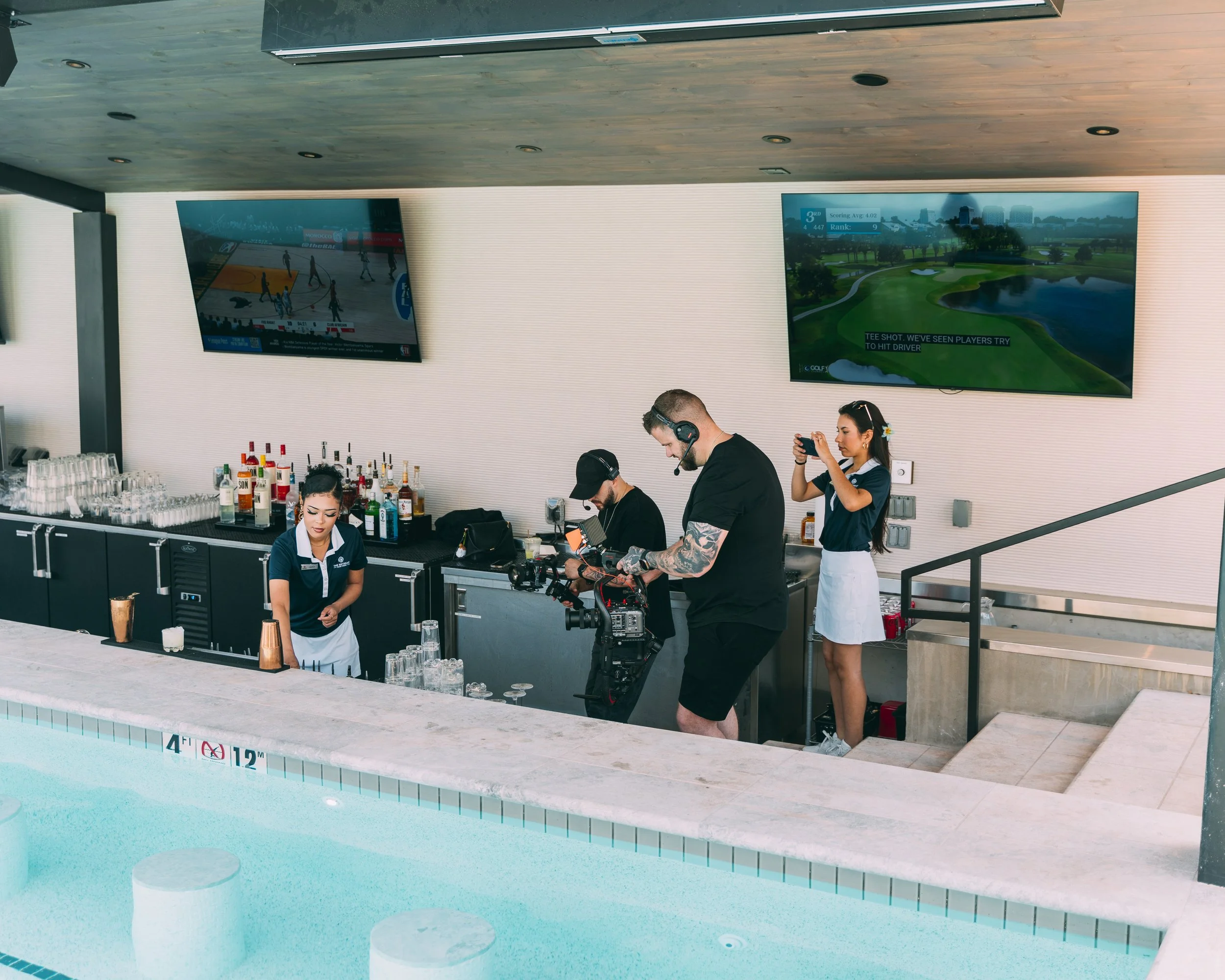 Poolside bar with bartenders and a cameraman recording. Two television screens display sports and a golf course. The pool has clear blue water and a depth marker of 4 feet 12 inches.