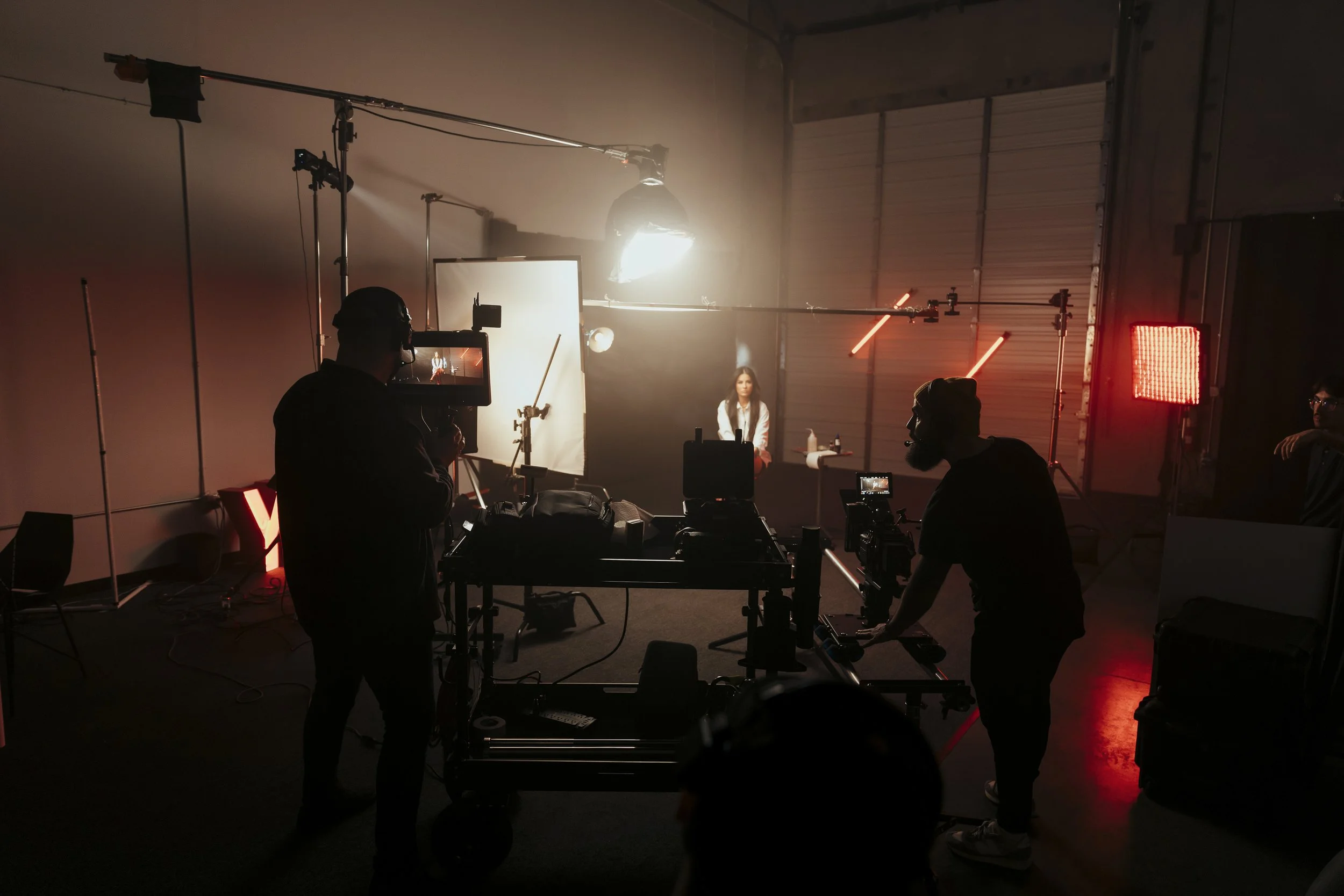 Behind the scenes of a video shoot with a woman sitting in front of a white backdrop, lighting equipment, and crew members operating cameras and equipment in a warehouse or studio setting.