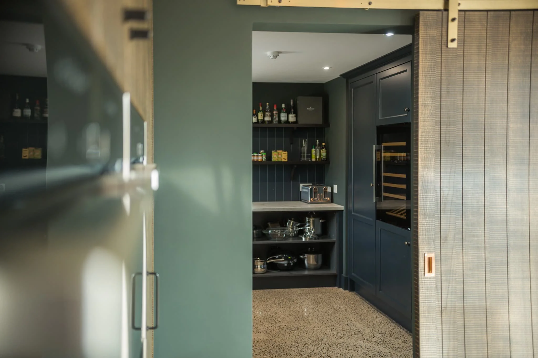 Looking through a sliding wooden doorway to a bespoke kitchen pantry with dark cabinets and built in wine fridge - part of Darren Morgan Design's bespoke kitchen design service