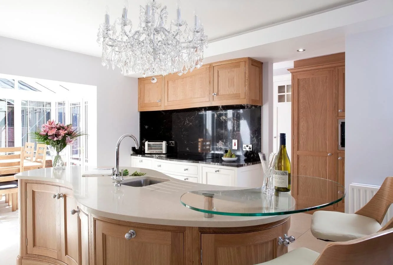A luxury traditional kitchen renovation with wooden cabinets and a kitchen island with glass extensions, white worksurface and a sink in the middle. To the left is a conservatory room, behind is another work surface with a black backsplash