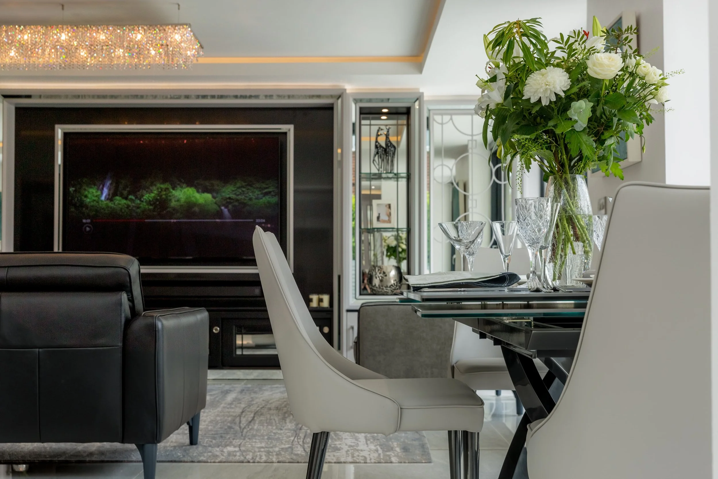 A living room with a black leather sofa facing a large entertainment system on a black wall, to the right is a glass dining table with white leather chairs around it and a large bouquet of white flowers and greenery in the middle