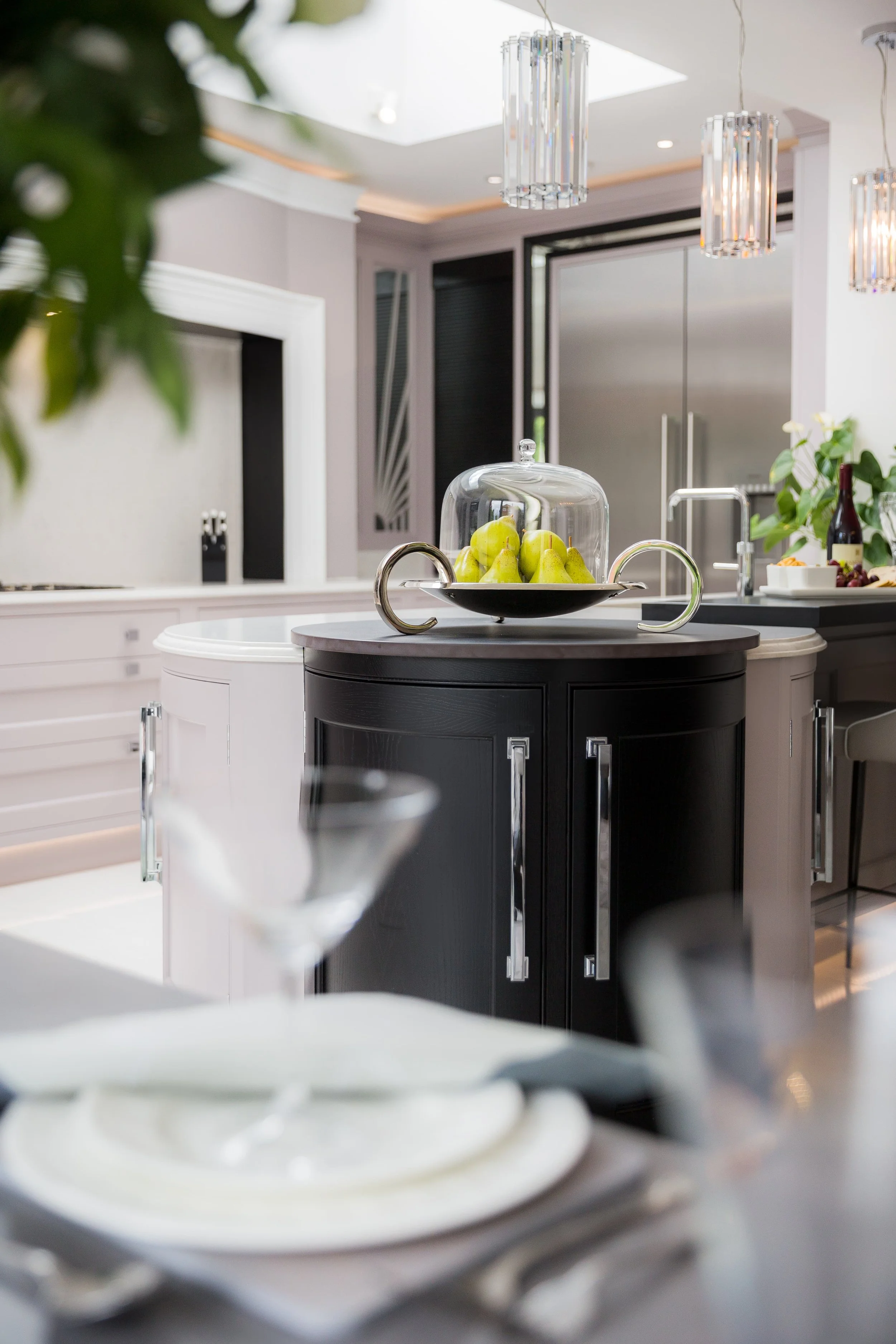 high end bespoke kitchen design with bespoke black and cream kitchen island cabinets in the centre of the room with a fruit display on the surface. In the background is a large double fridge, in the foreground blurred is glassware and cutlery