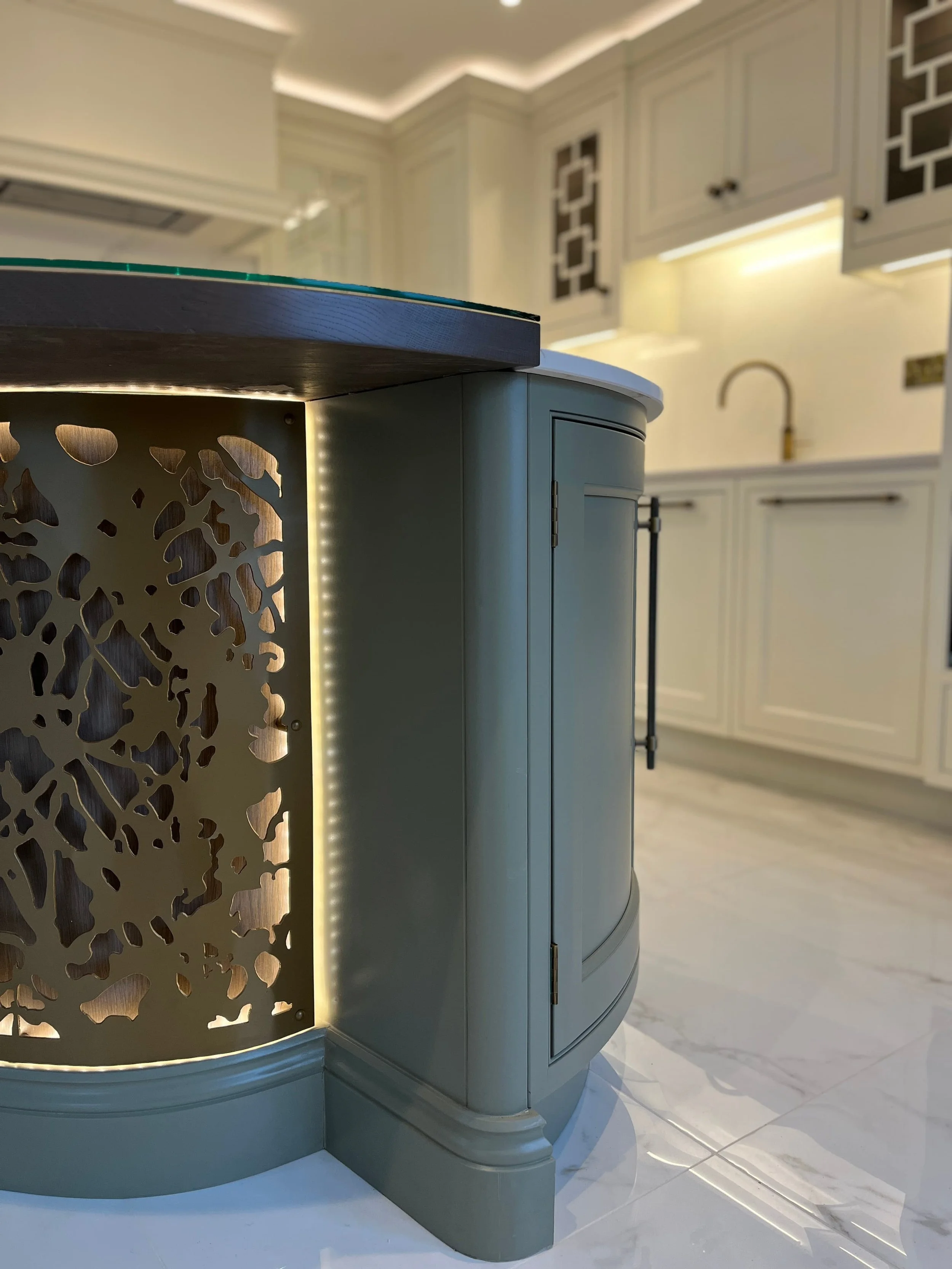 A close up of a dark grey-green kitchen island cabinet, metal overlay in backlit under the dining surface and the rest of the kitchen seen in the foreground is cream - kitchen remodel by online kitchen designer Darren Morgan