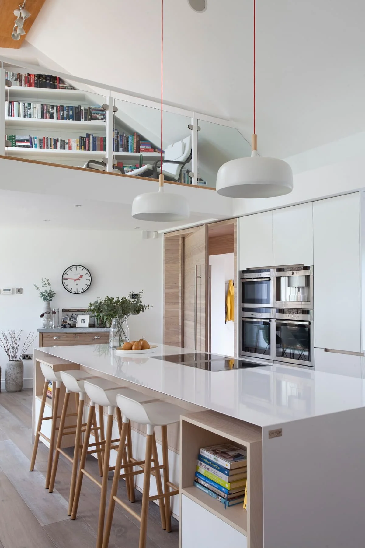 A white minimalist kitchen with an island in the middle and four stools tucked underneath. A double oven on the far wall with light wood accents on the door, a chest of draws and stool legs. Vaulted ceiling and glass balcony partially shows a library