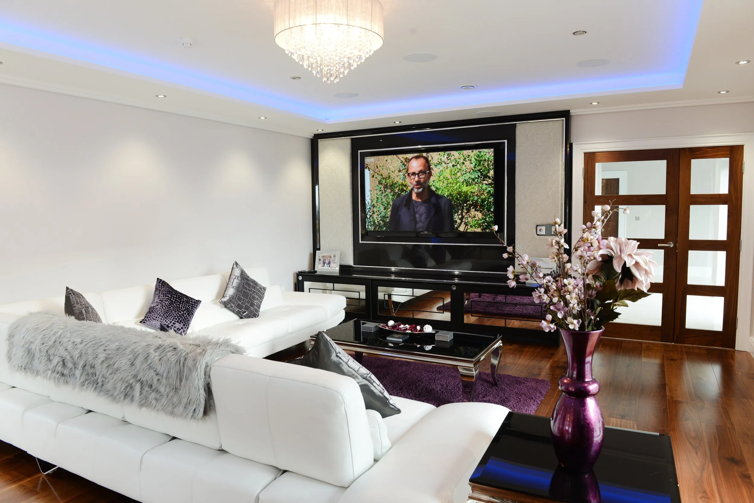 a white room with a white  L-shaped leather sofa facing a large tv and home entertainment system mounted on a mirrored wall