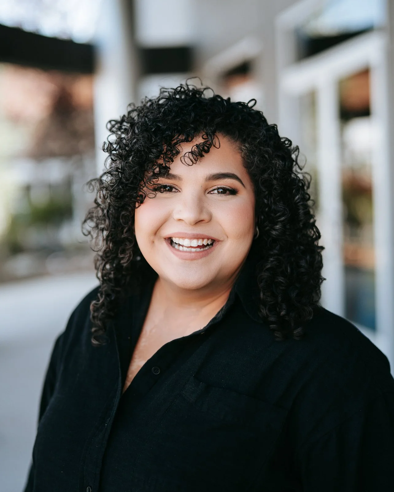 A woman with dark curly hair smiling and standing in an indoor space with large windows and bright lighting, wearing a black top and a dark checked blazer.