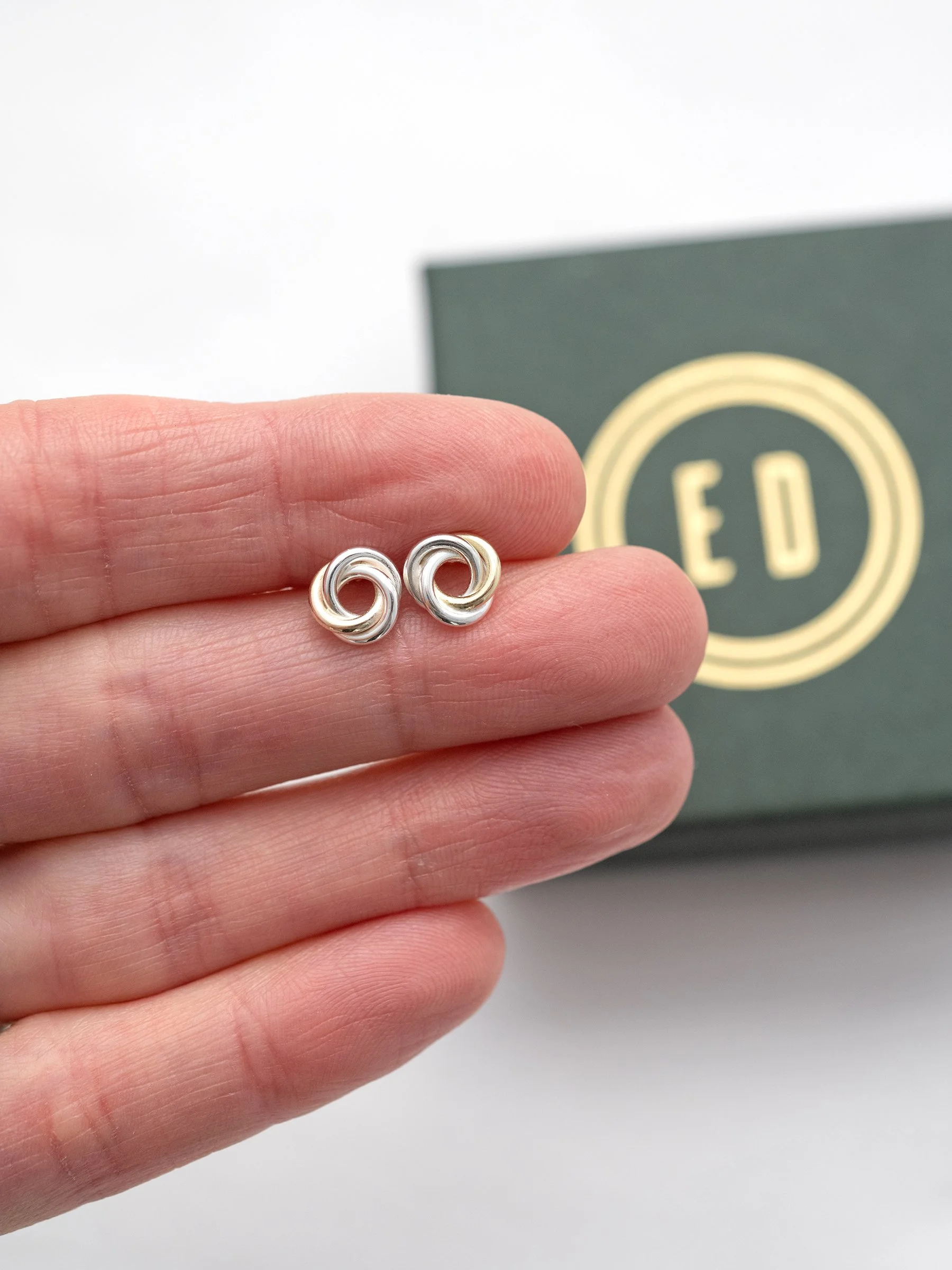 A pair of handmade gold and silver Russian ring stud earrings displayed in a persons hand with a jewellery gift box in the background.