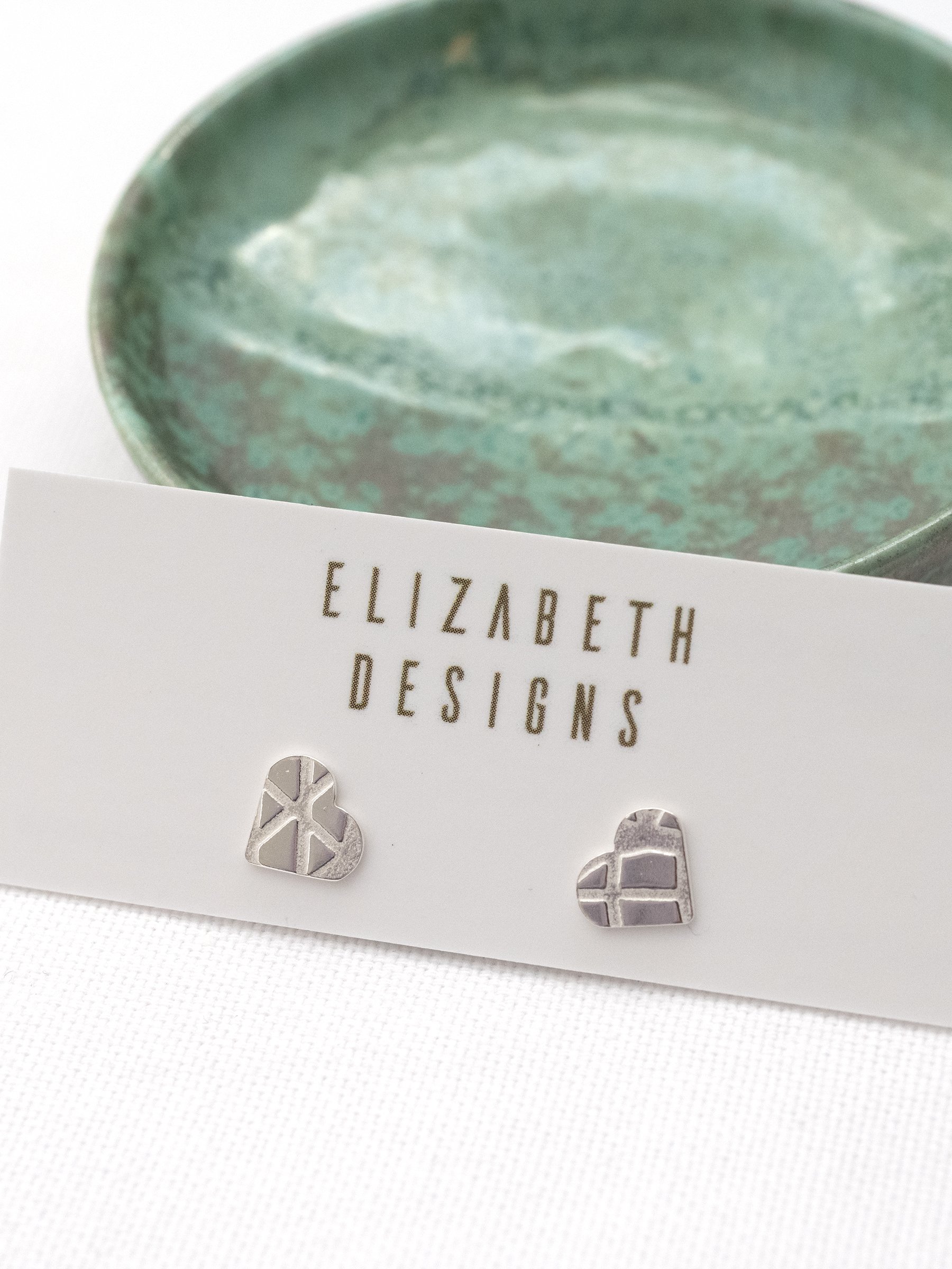 A pair of silver pattern heart stud earrings displayed on a jewellery display card with a handmade green jewellery dish in the background.