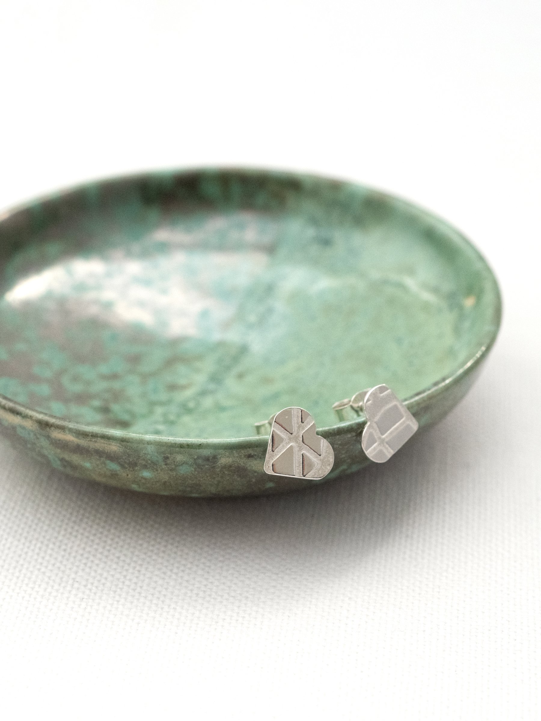 A pair of silver patterned heart stud earrings displayed on the edge of a handmade green jewellery trinket dish.