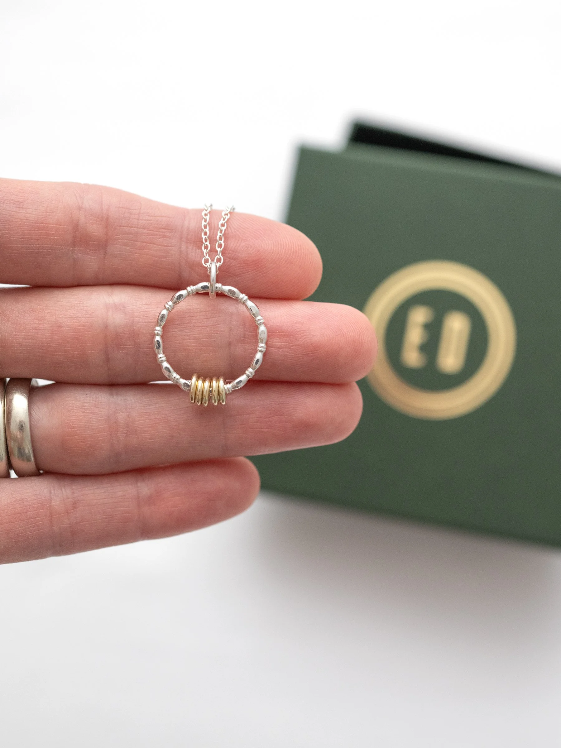 A person displaying a mixed metal 5 ring necklace in their hand with a green presentation box in the background.
