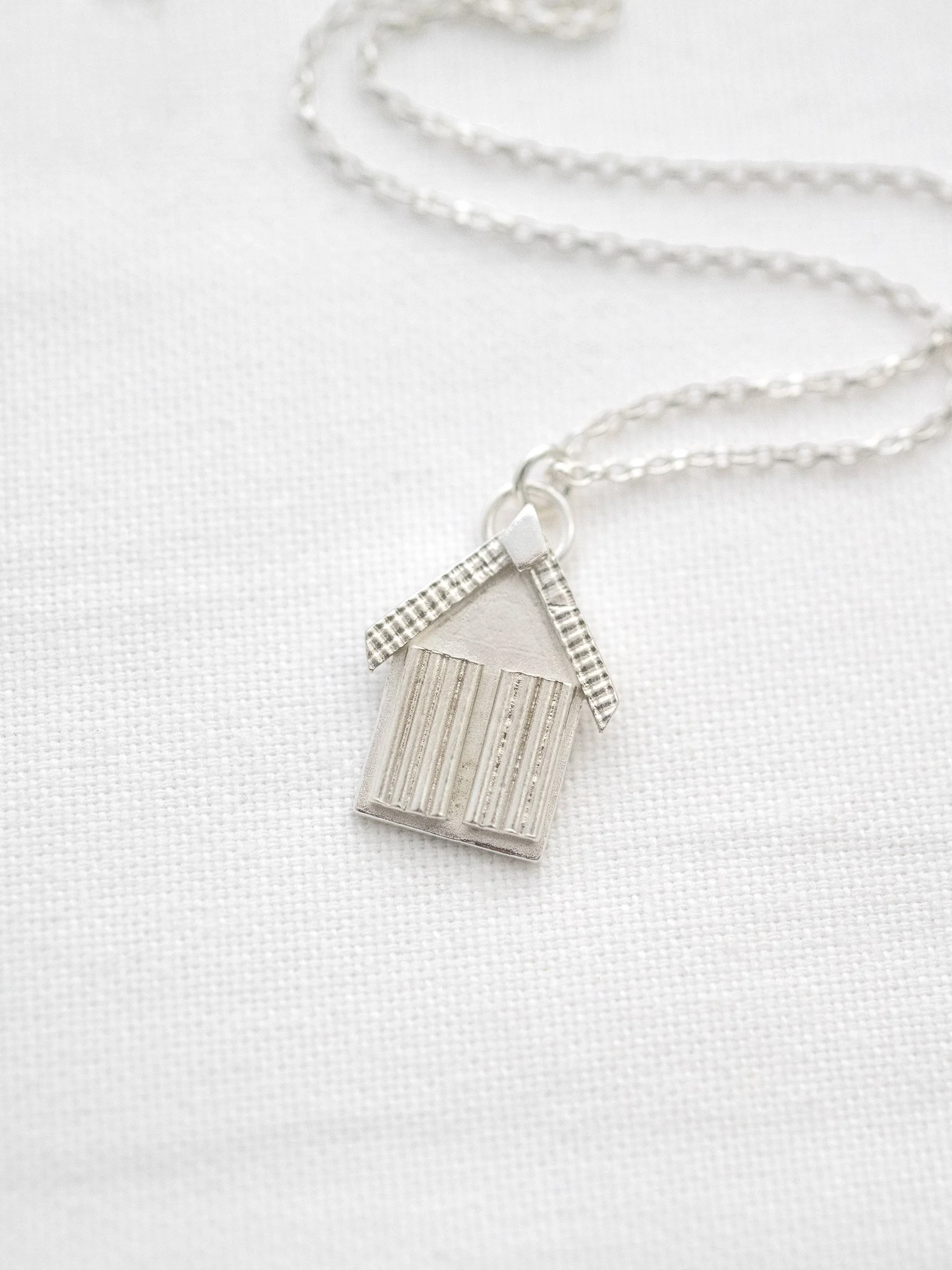 A silver beach hut necklace displayed on white material.