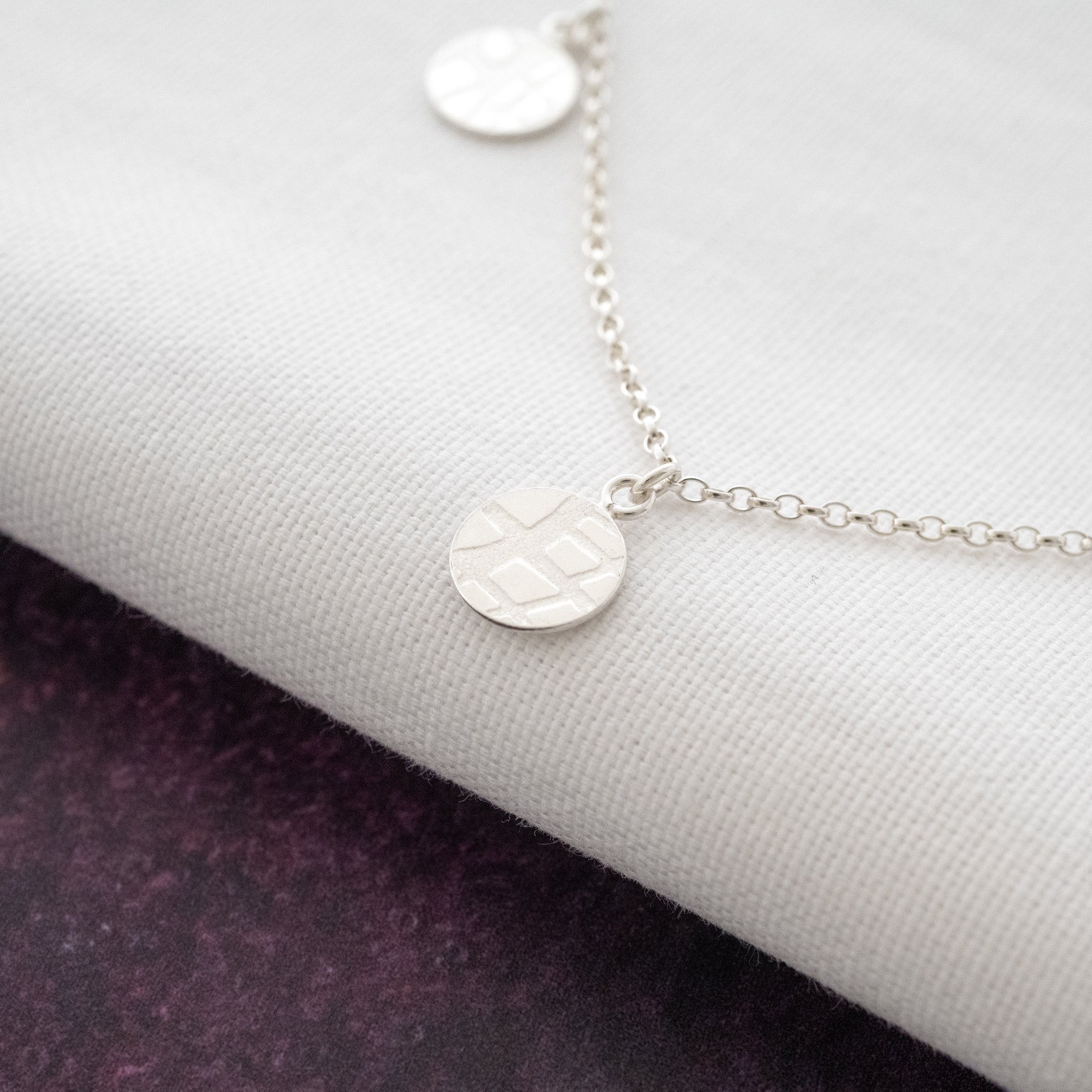 A side view of a silver textured disc necklace displayed over a white and purple background.