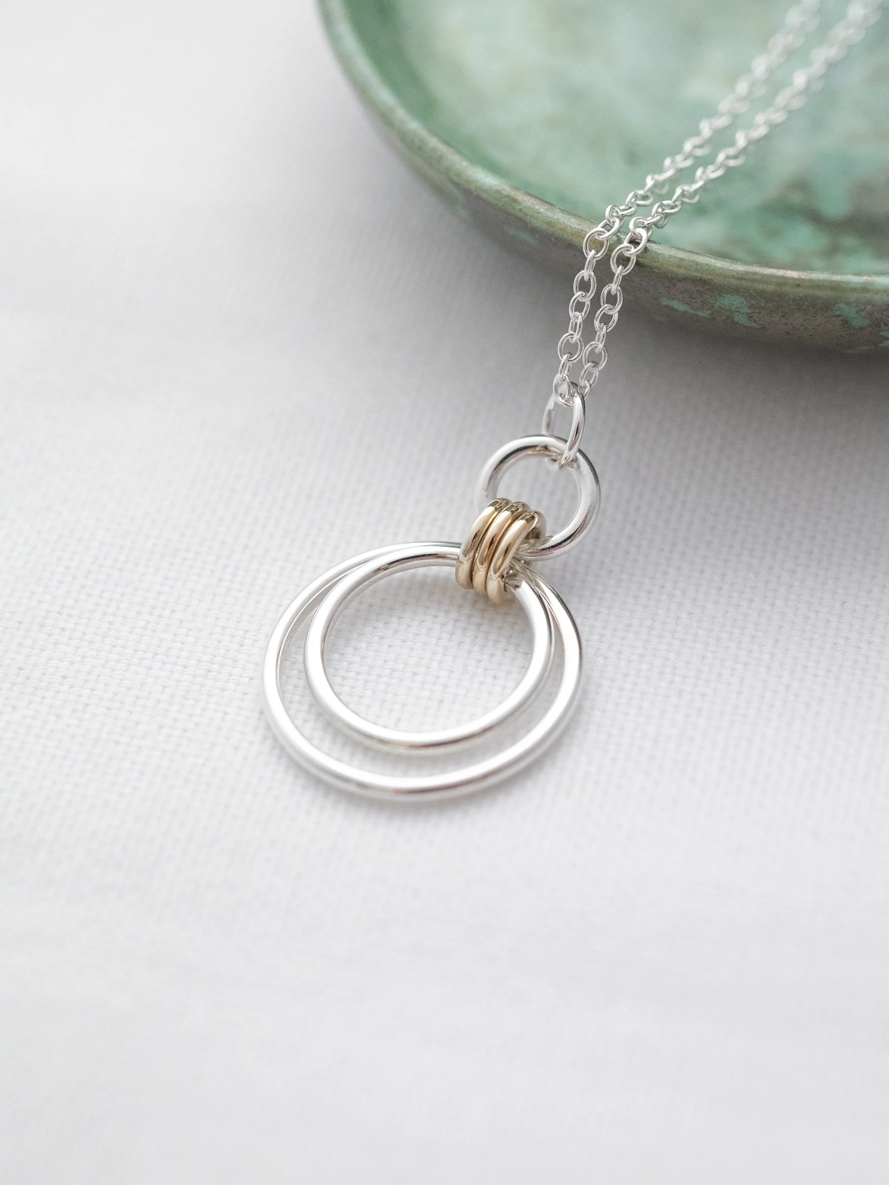 A close up of a gold and silver circle necklace displayed on white material with a green jewellery dish in the background.
