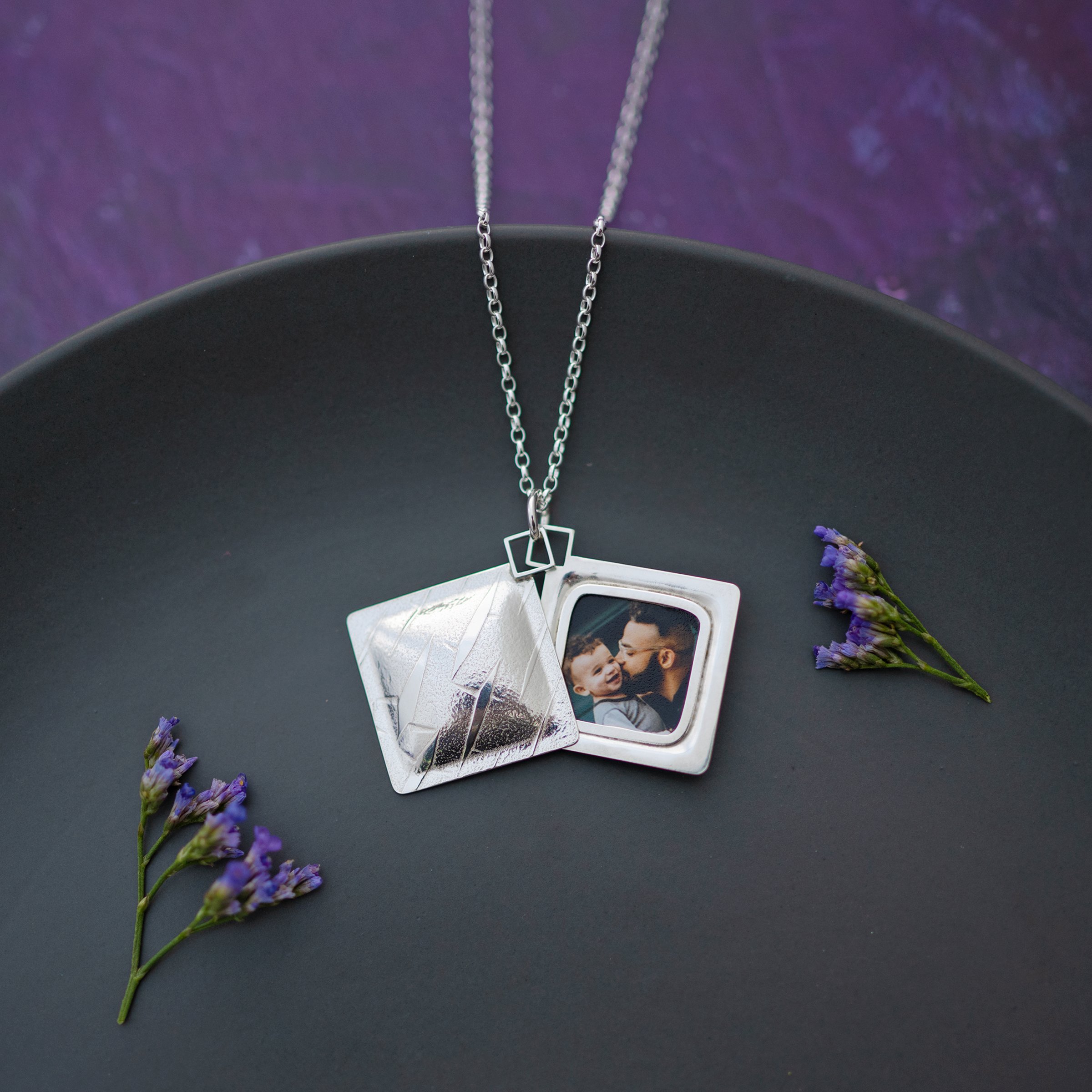 An open, square diamond shaped locket displaying a photo inside against a grey and purple background.