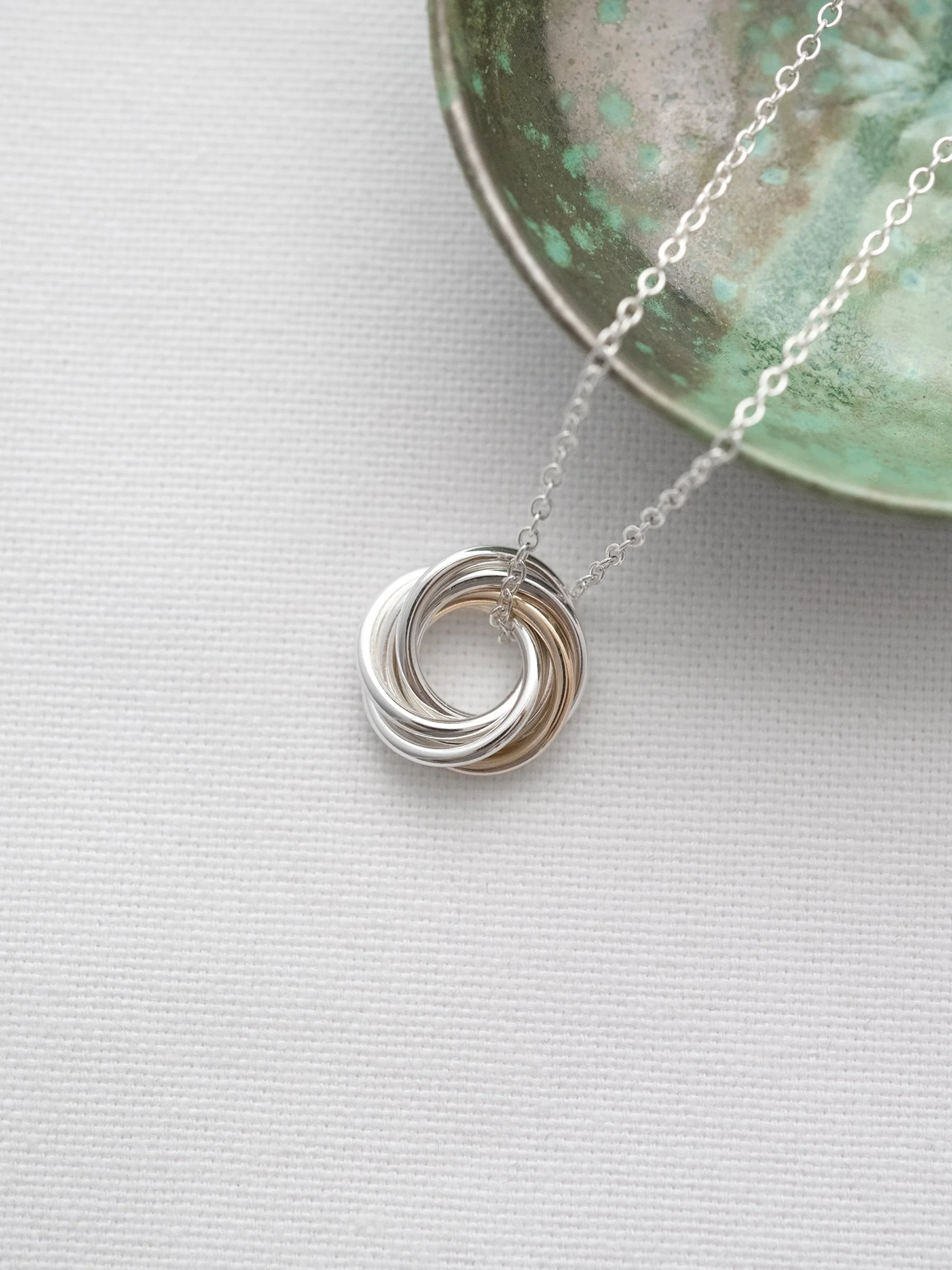 A gold and silver 6 ring necklace for a 60th birthday displayed on white material with a green jewellery dish in the background.