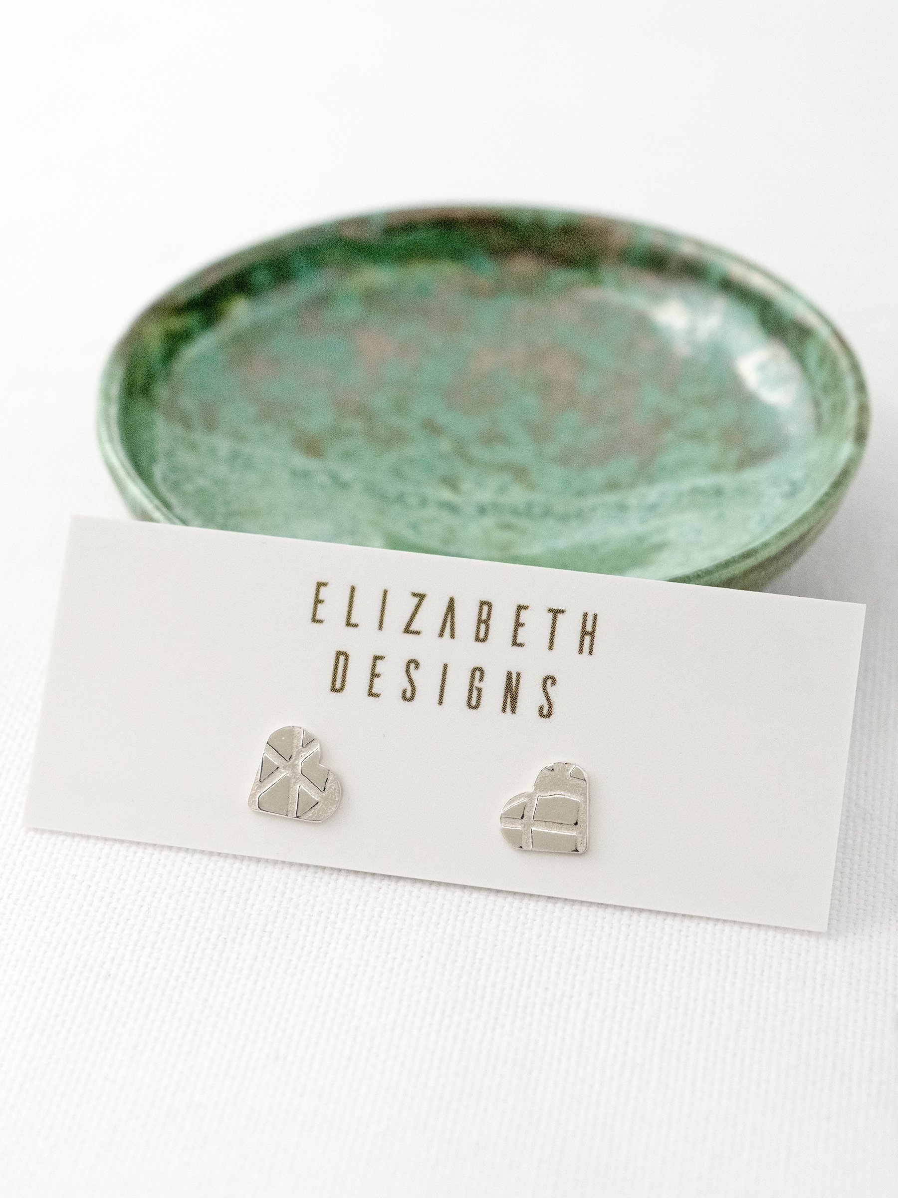 A pair of silver heart stud earrings displayed on a jewellery presentation card with a green jewellery dish in the background.
