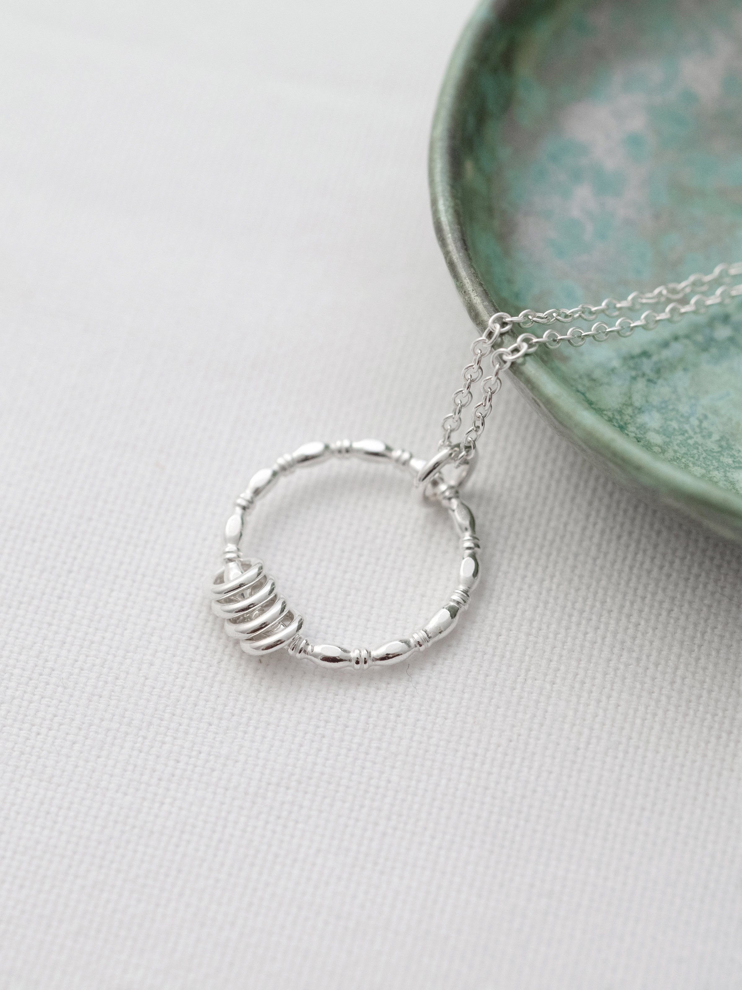 A side view of a silver 40th birthday ring necklace displayed over white material with a turquoise jewellery dish in the background.