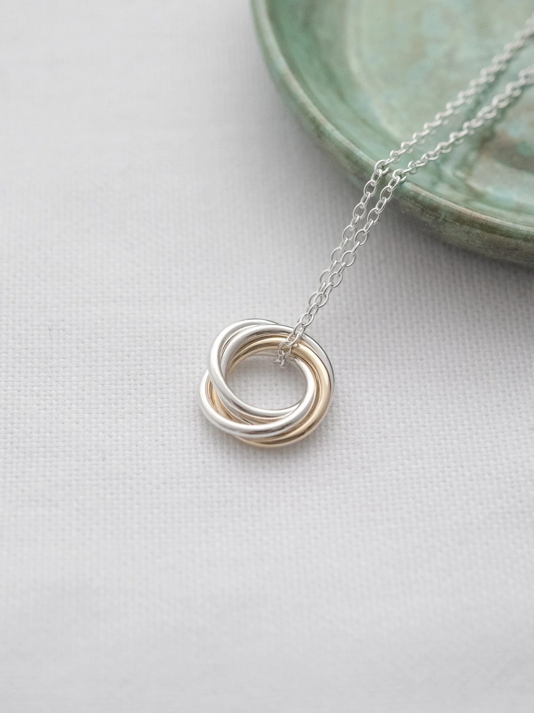 A unique 4 ring gold and silver necklace displayed on white material with a green jewellery dish in the background.