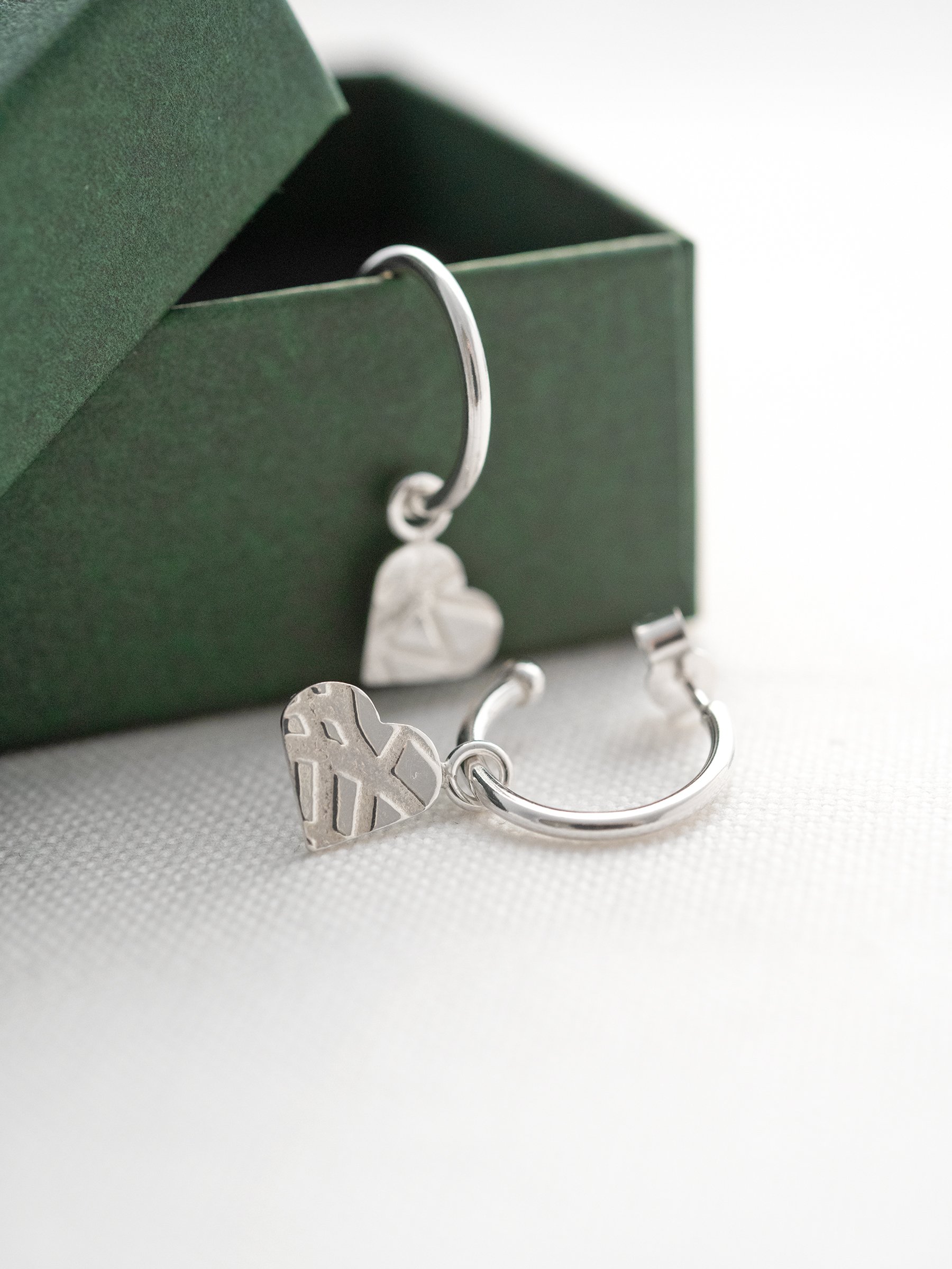 Sterling silver heart charm hoop stud earring displayed over a green jewellery presentation box.