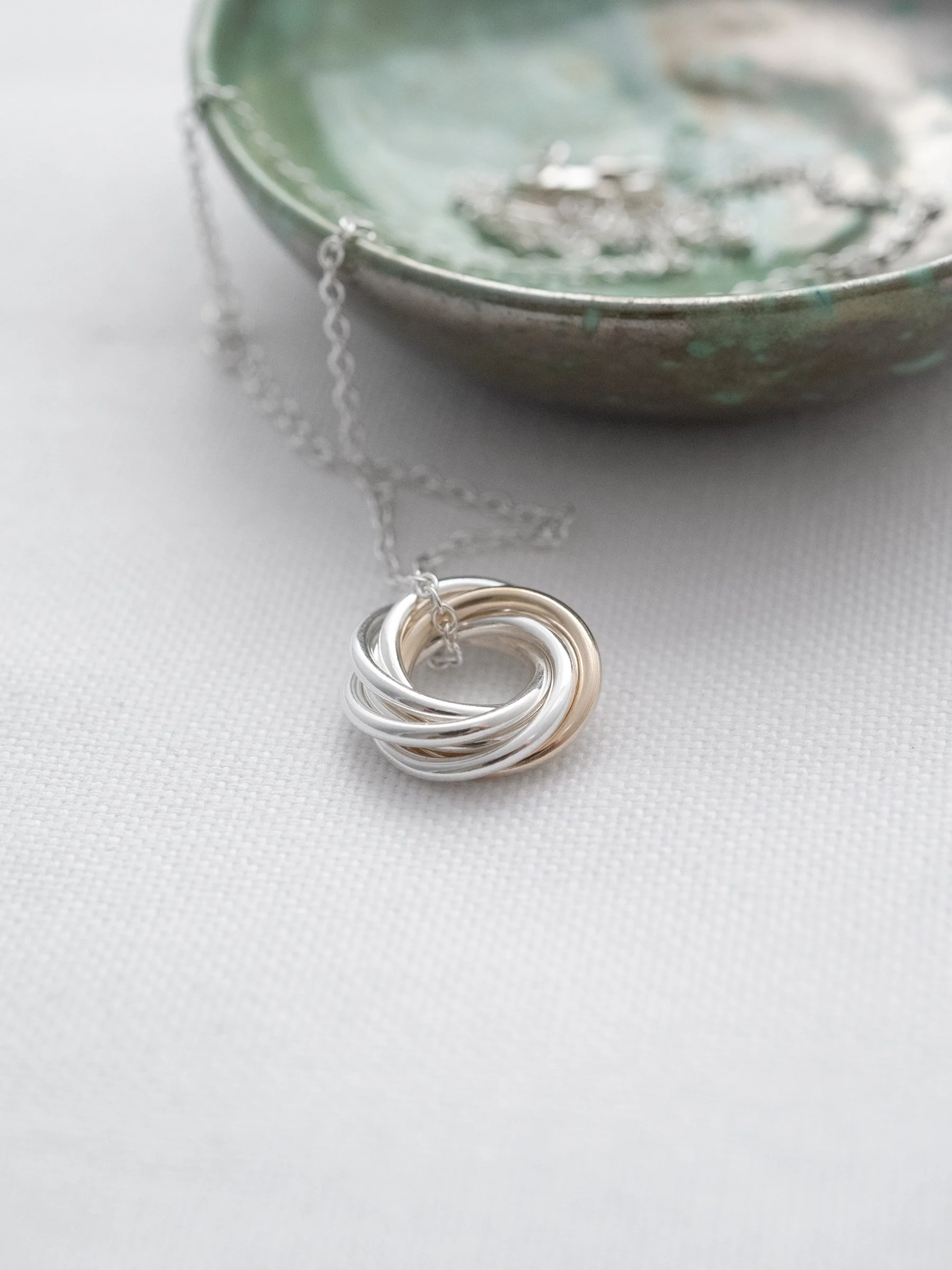 A close up of a mixed metal ring necklace displayed on white material with a green jewellery dish in the background.