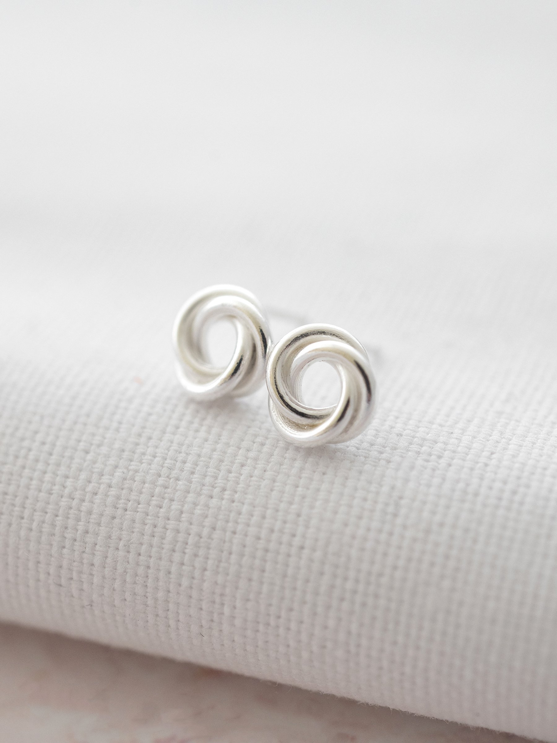 A pair of sterling silver knot stud earrings displayed over white material against a pale pink background.