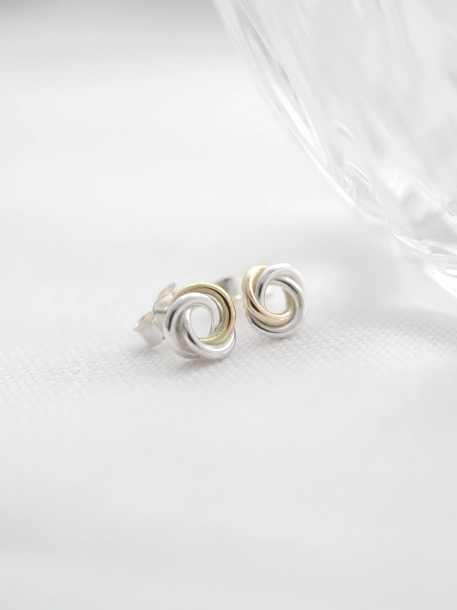 A side view of a pair of gold and silver circle stud earrings displayed on white material next to a decorative glass dish.