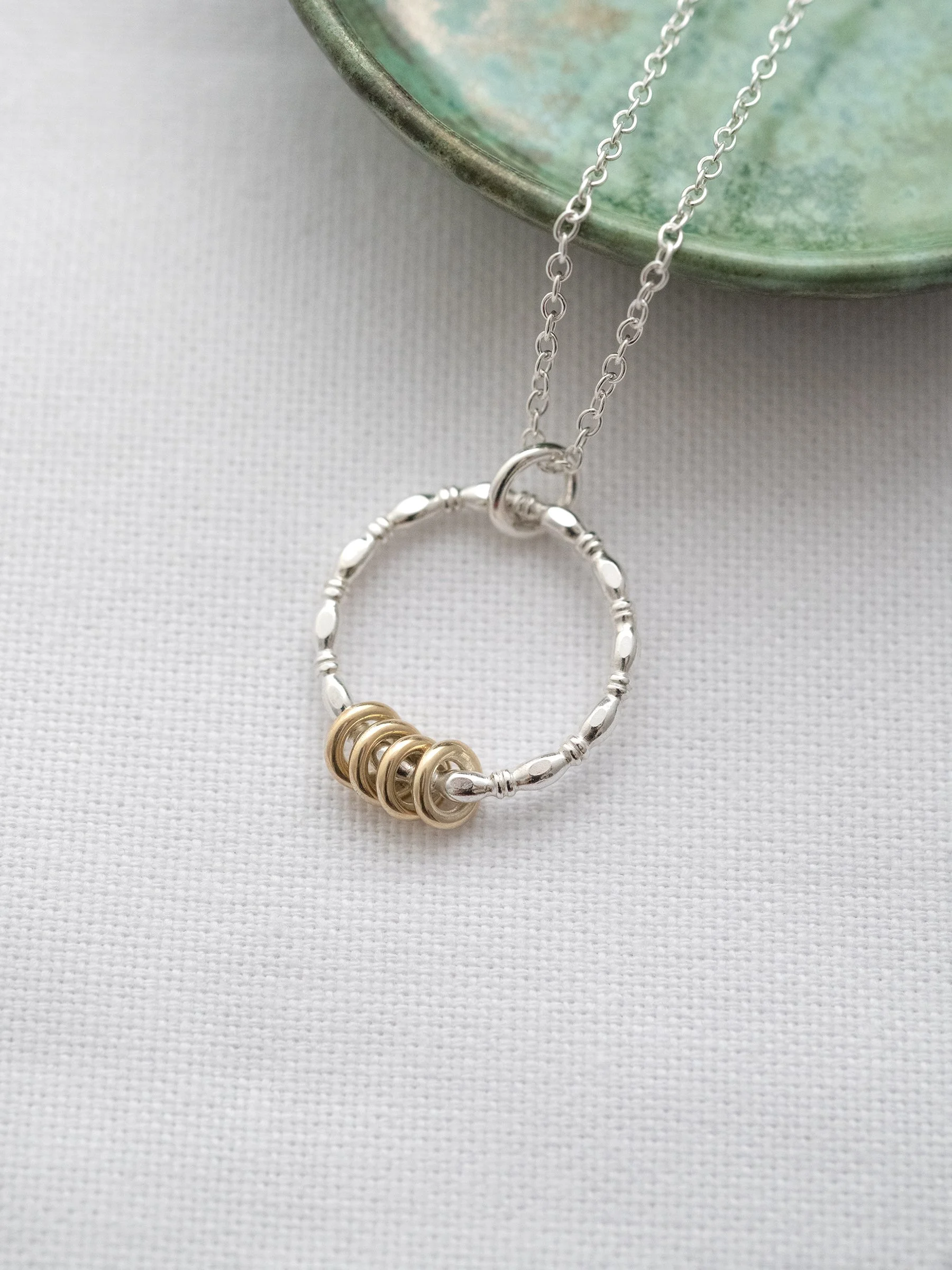 A gold and silver four ring necklace displayed on white material and a green jewellery dish in the background.