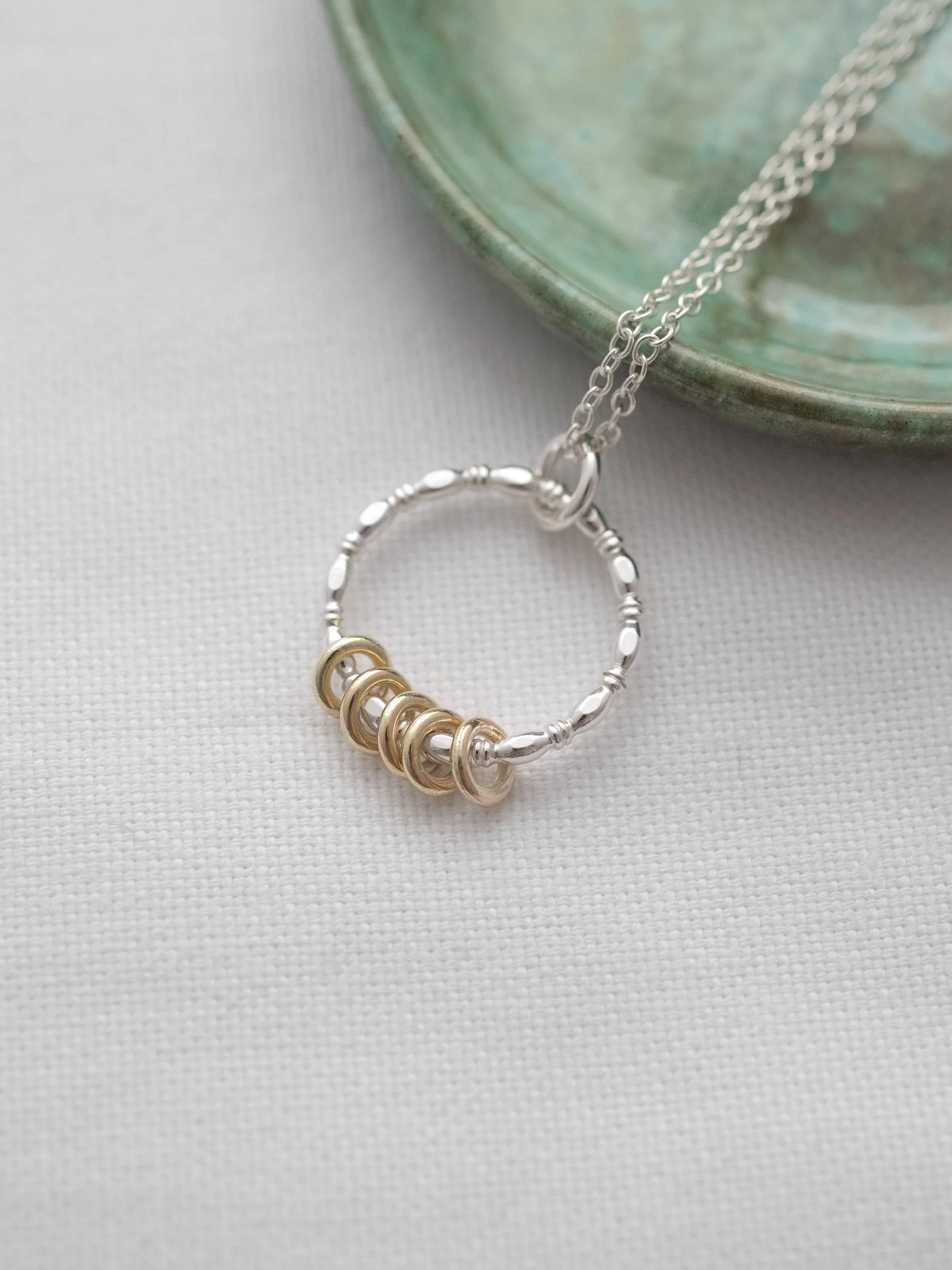 A close up of a silver necklace with 5 gold rings for a 50th birthday displayed next to a green jewellery dish on white material.