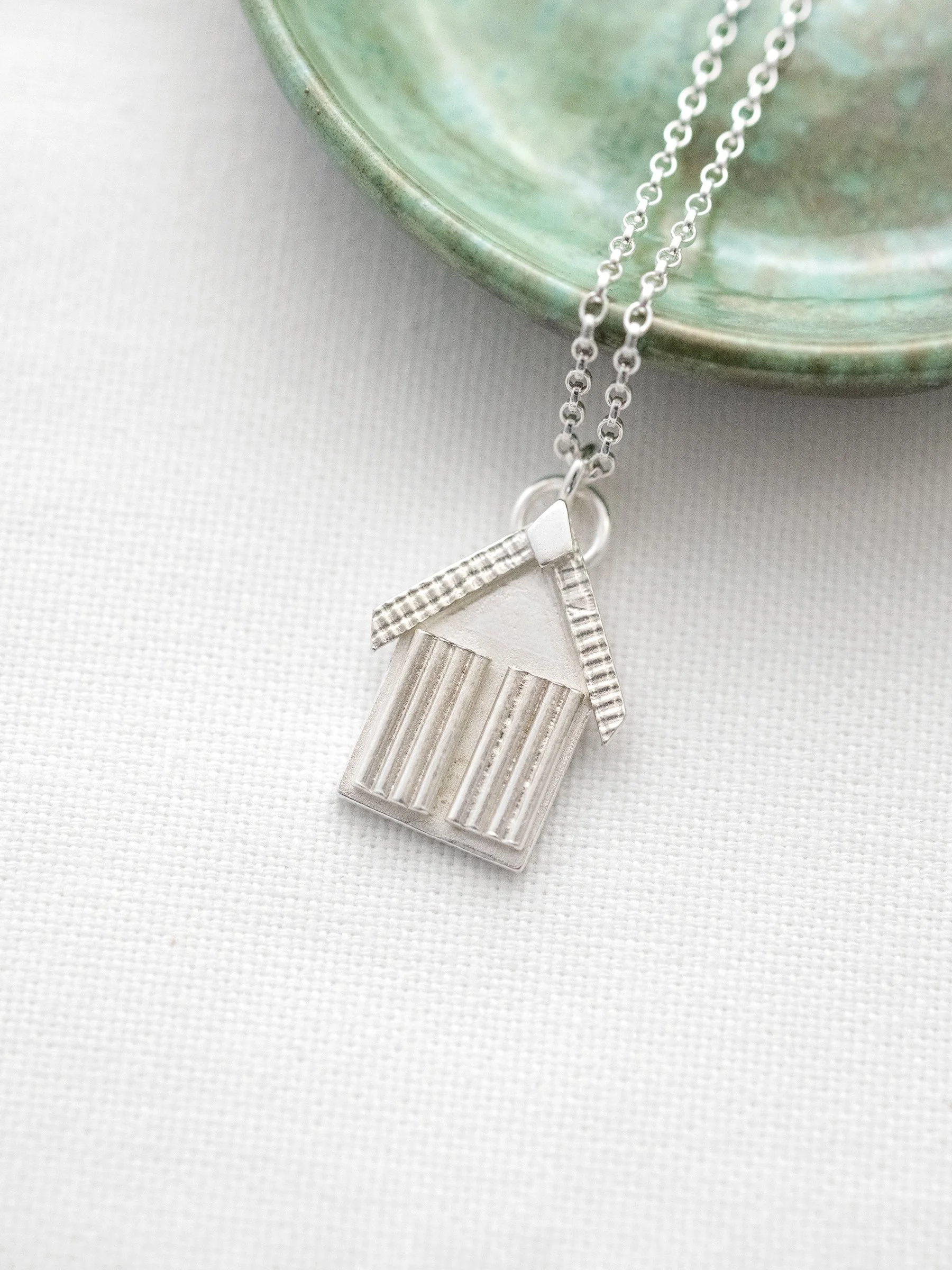 A unique sterling silver beach hut necklace displayed next to a green jewellery dish.