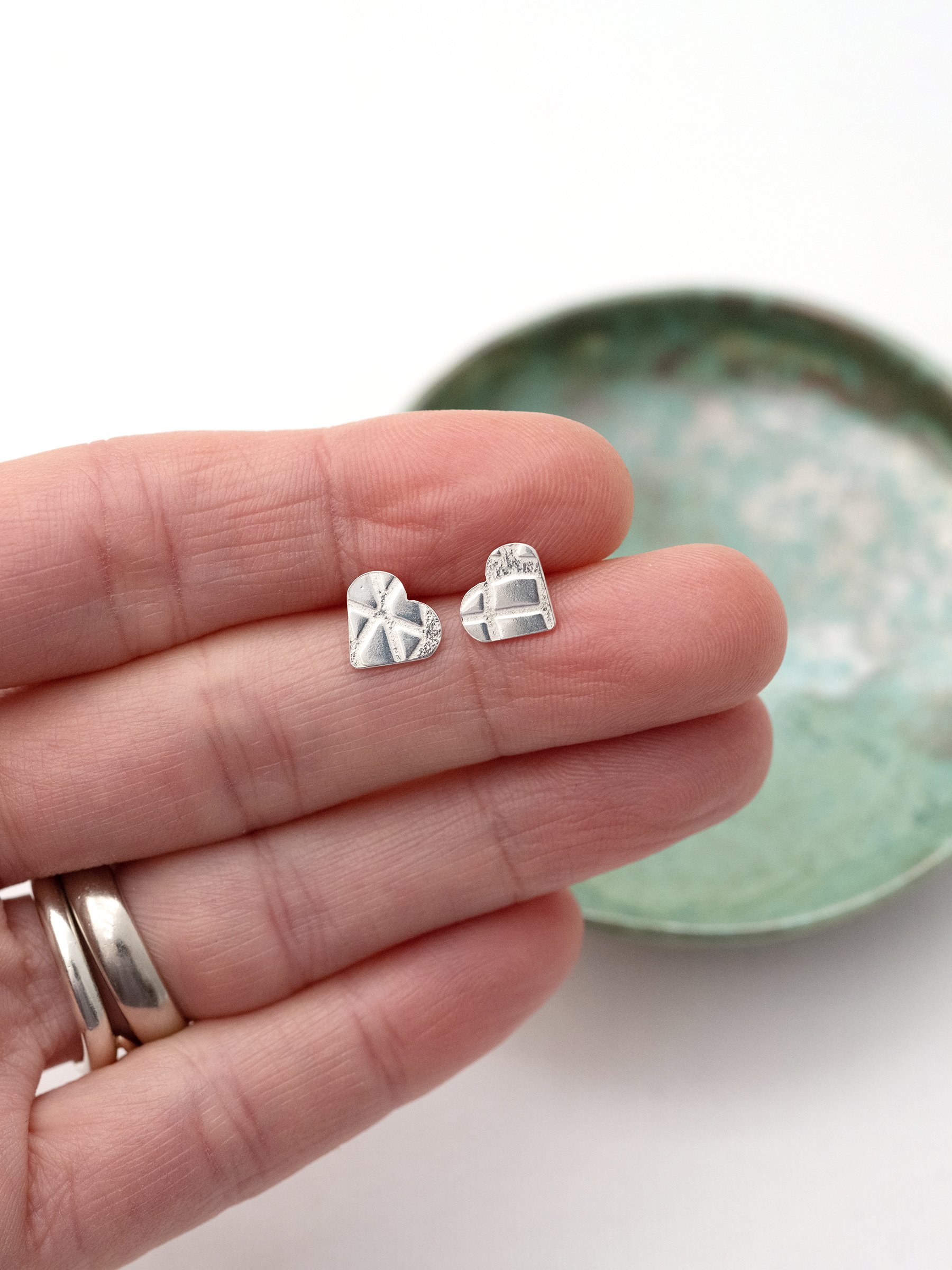 A pair of sterling silver heart stud earrings displayed on a persons hand with a handmade jewellery green dish in the background.