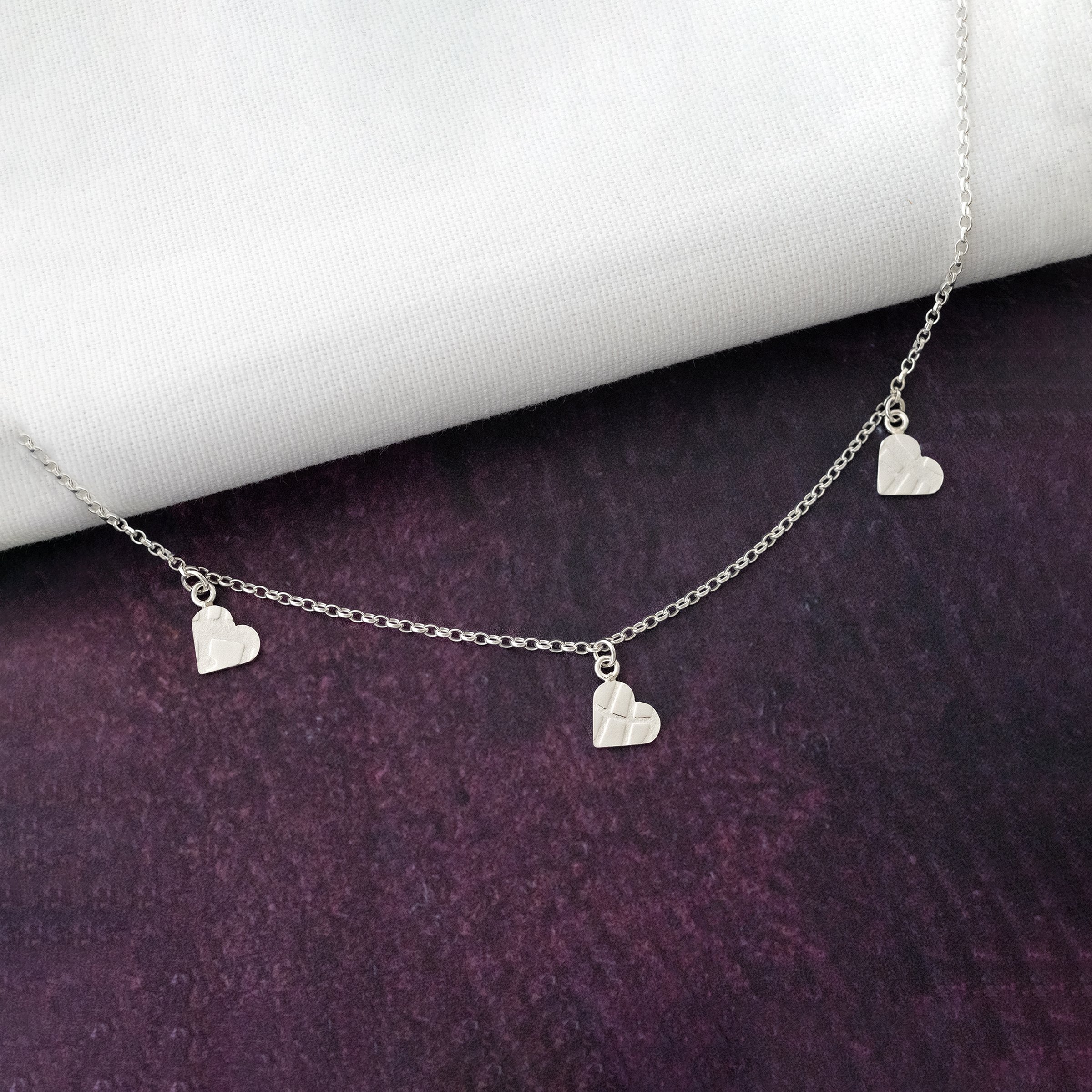 A silver heart textured necklace displayed over white material and a purple background.