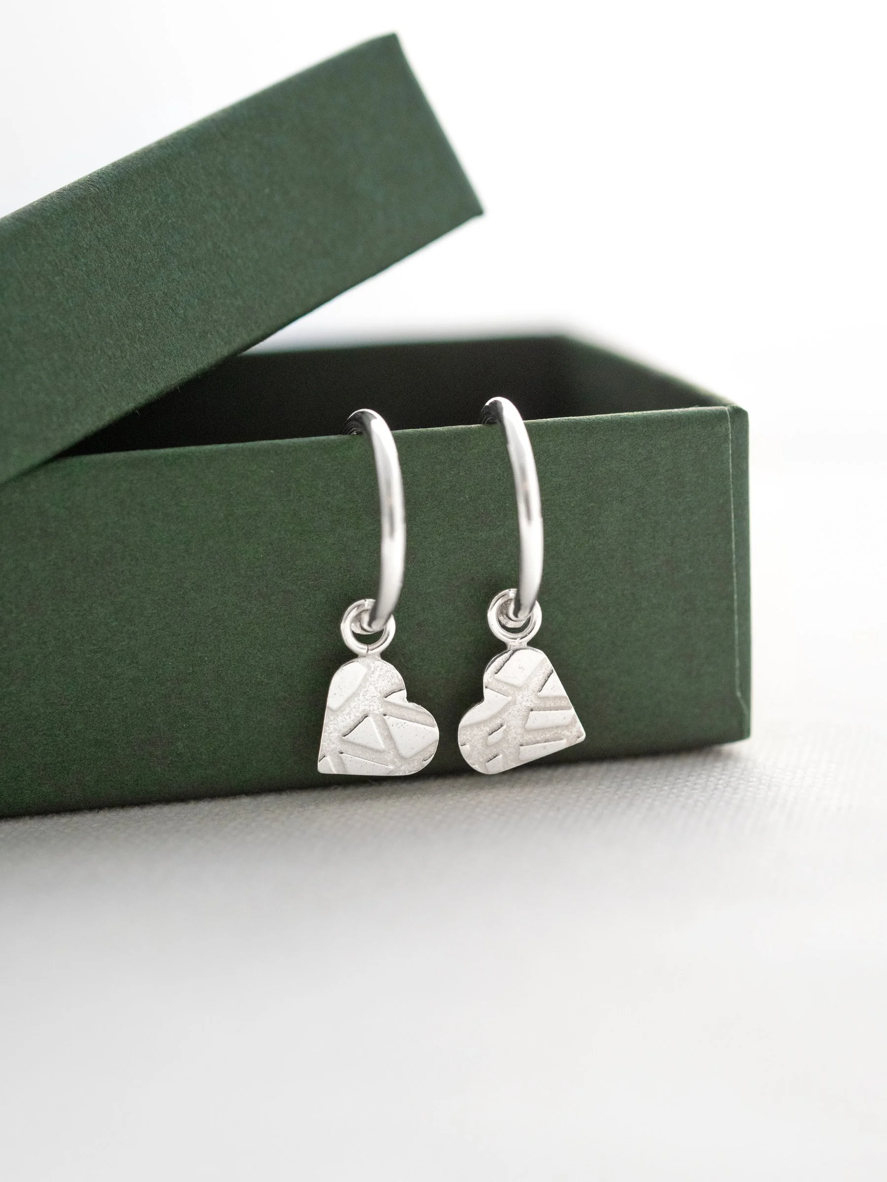 A pair of patterned heart hoop stud earrings hanging over a green jewellery presentation box.