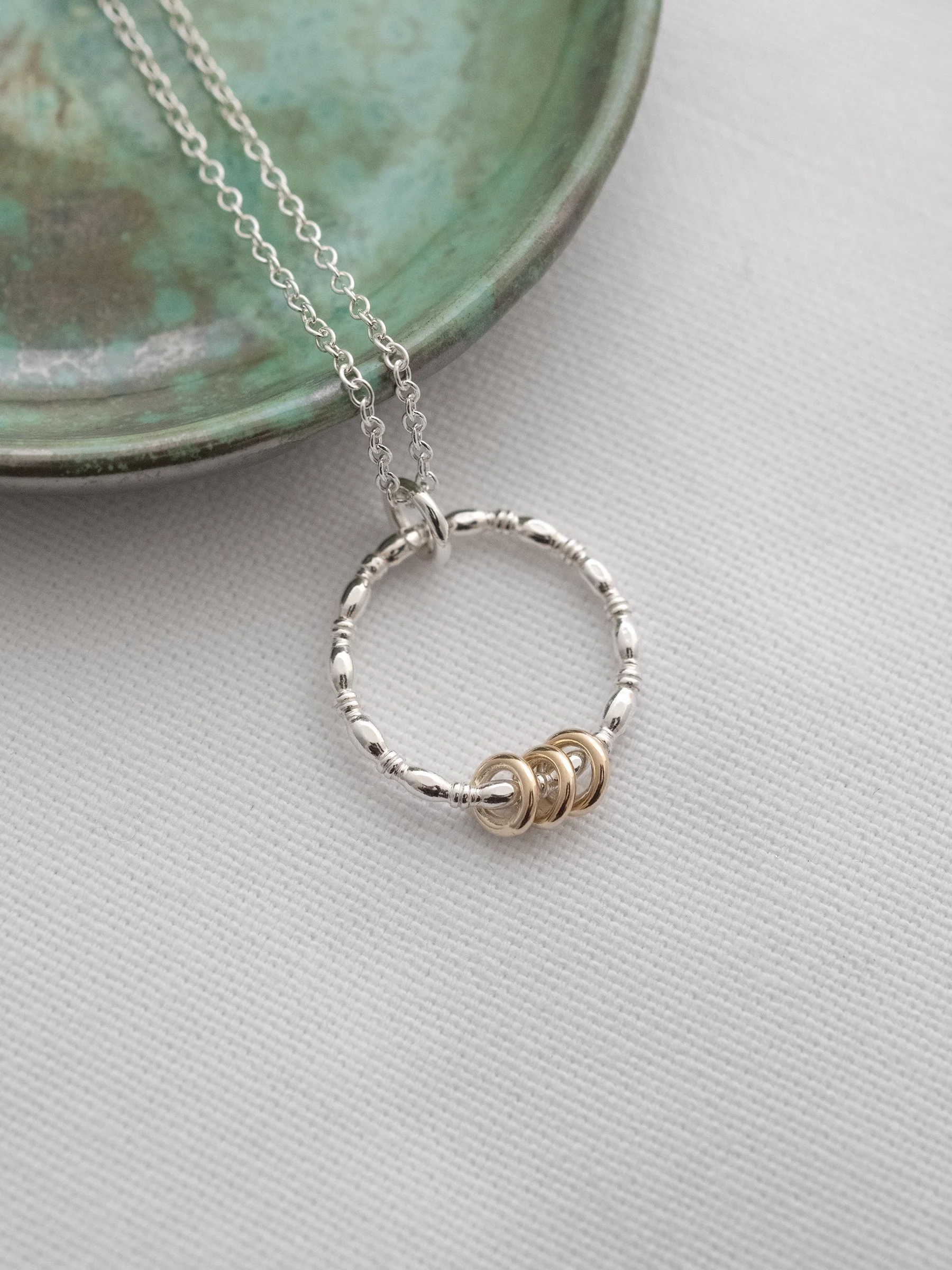 A sterling silver and gold 3 ring necklace for a 30th birthday displayed on white material with a green jewellery dish in the background.