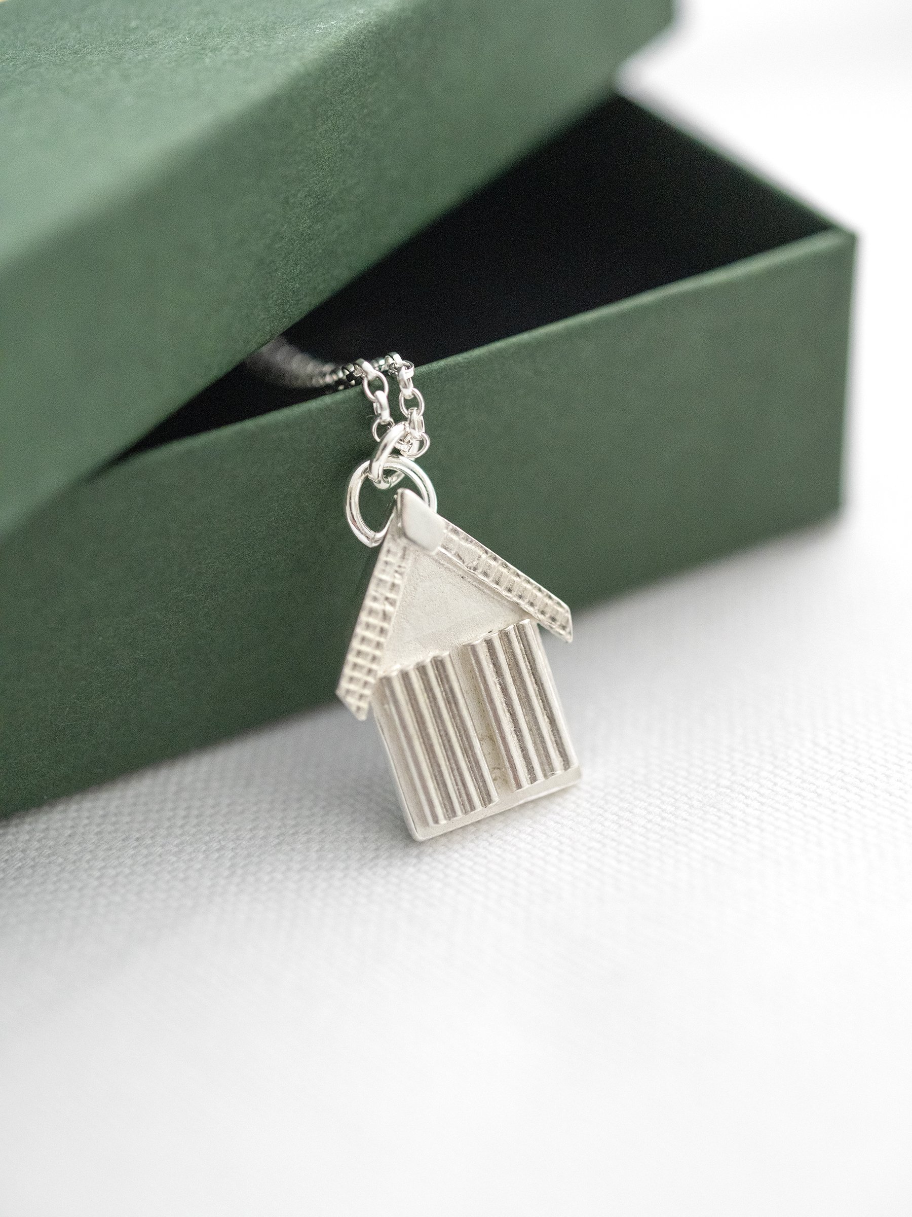 A side view of a silver beach hut pendant leaning against a green jewellery presentation box.