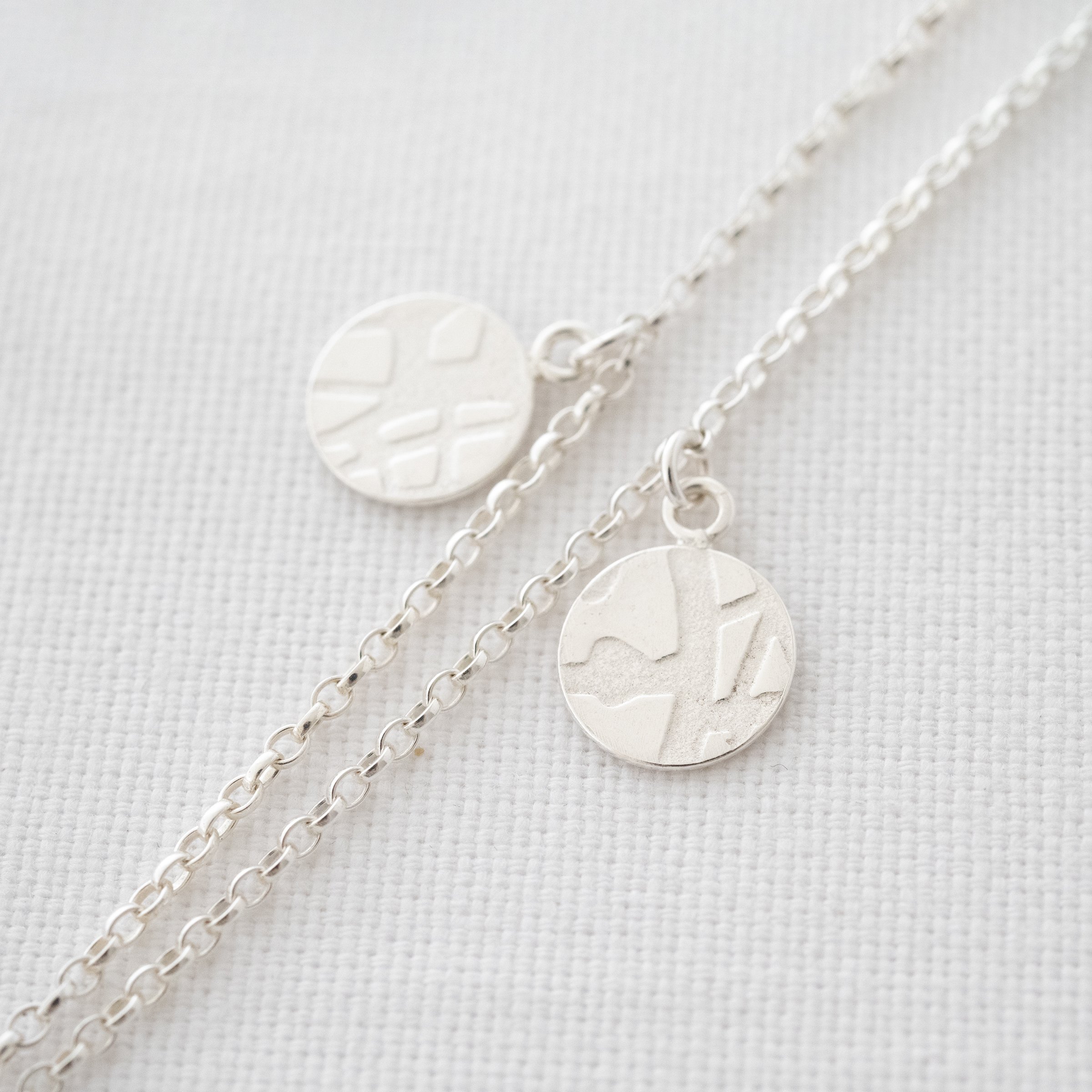 A close up of a silver textured disc necklace displayed against white material.