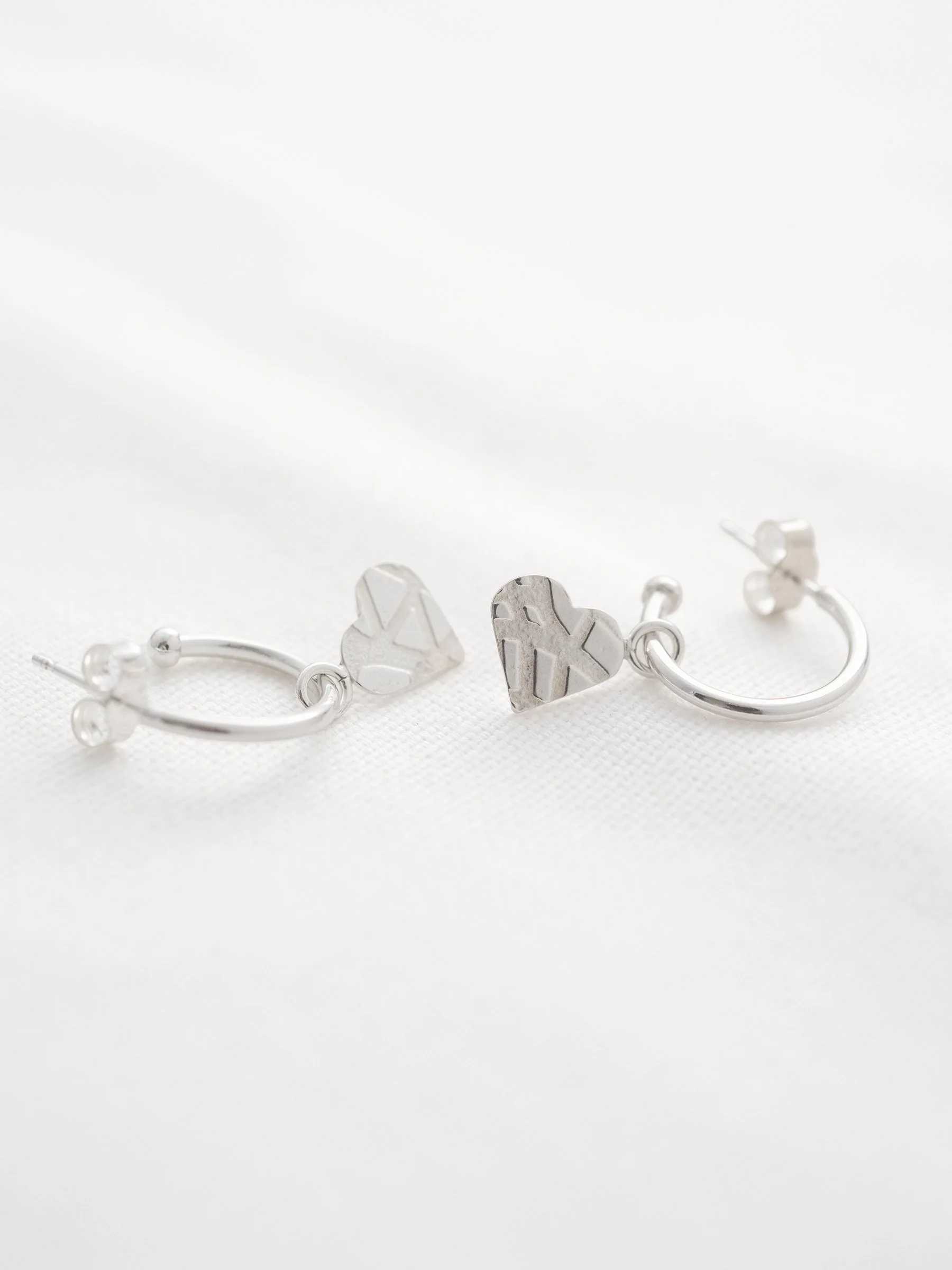 A pair of silver hoop stud earrings with patterned silver hearts displayed on white material.