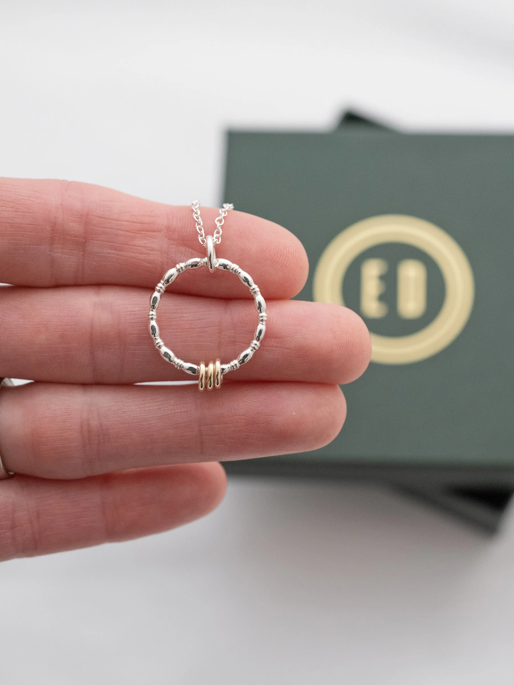 A silver and gold 3 ring necklace displayed in a persons hand with the jewellery presentation box in the background.