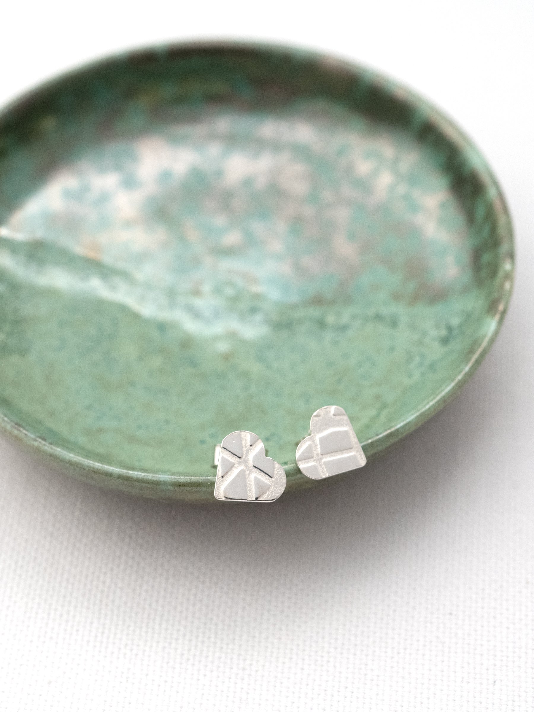 A pair of silver heart stud earrings displayed on a handmade green trinket jewellery dish.