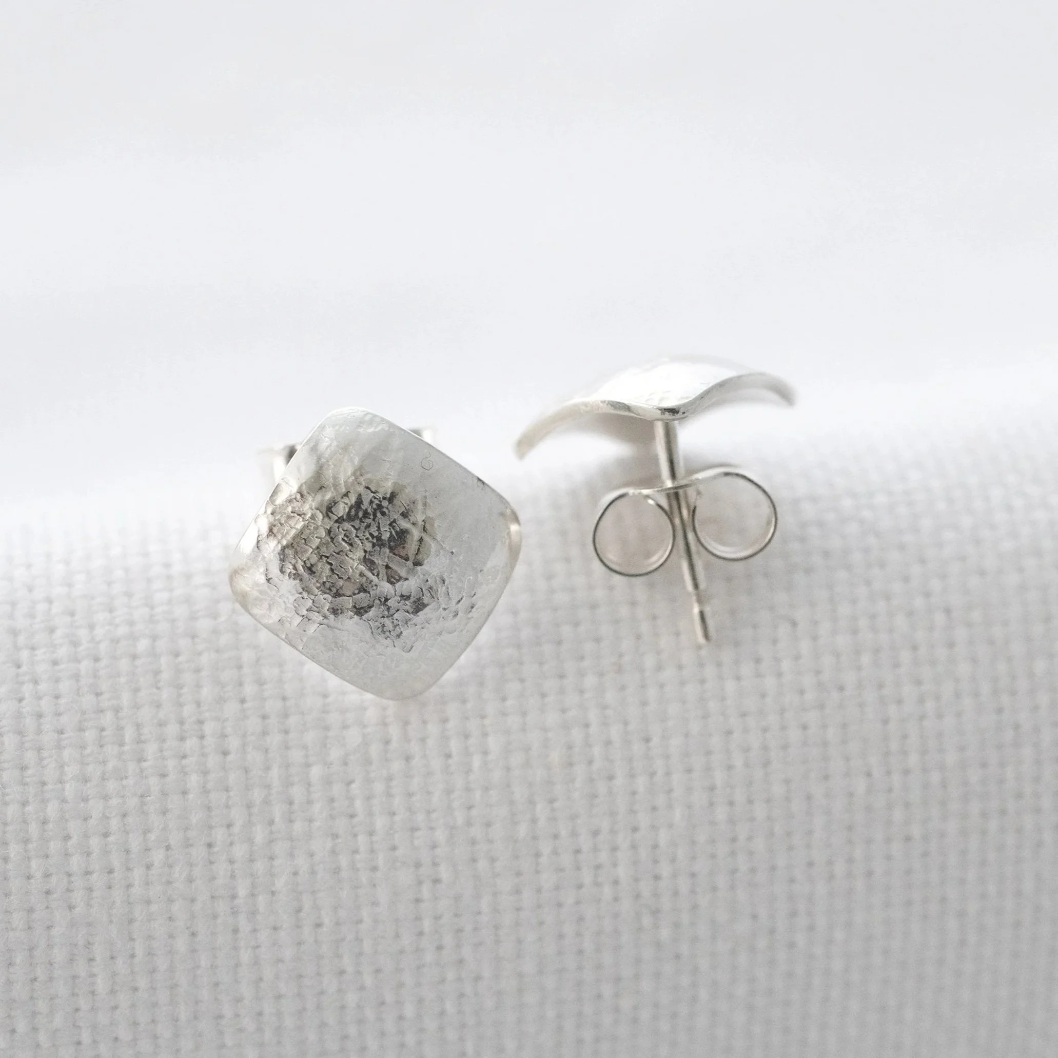 A close up of a pair of sterling silver texture square stud earrings displayed on white material.