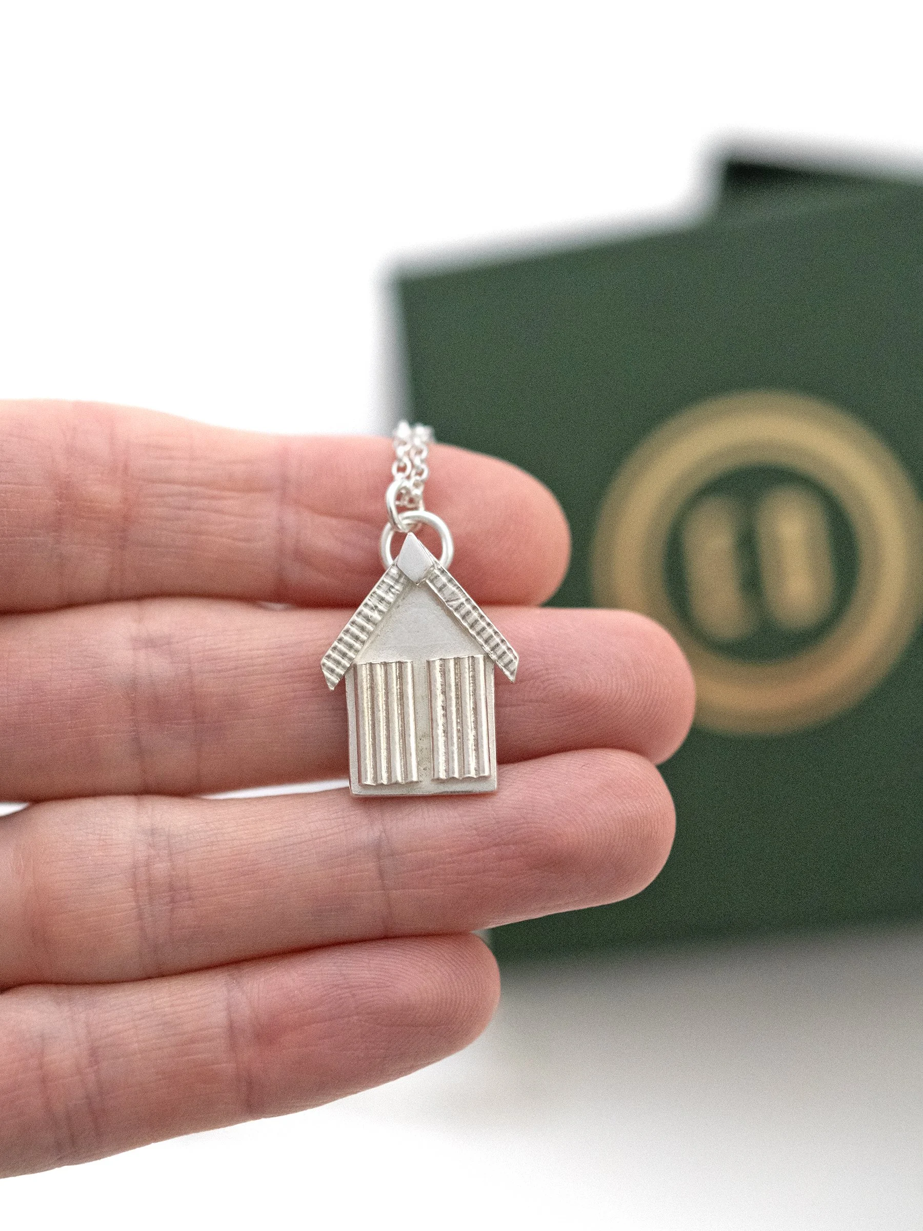 A close up of a silver beach hut necklace displayed in a persons hand with a green jewellery box in the back ground.