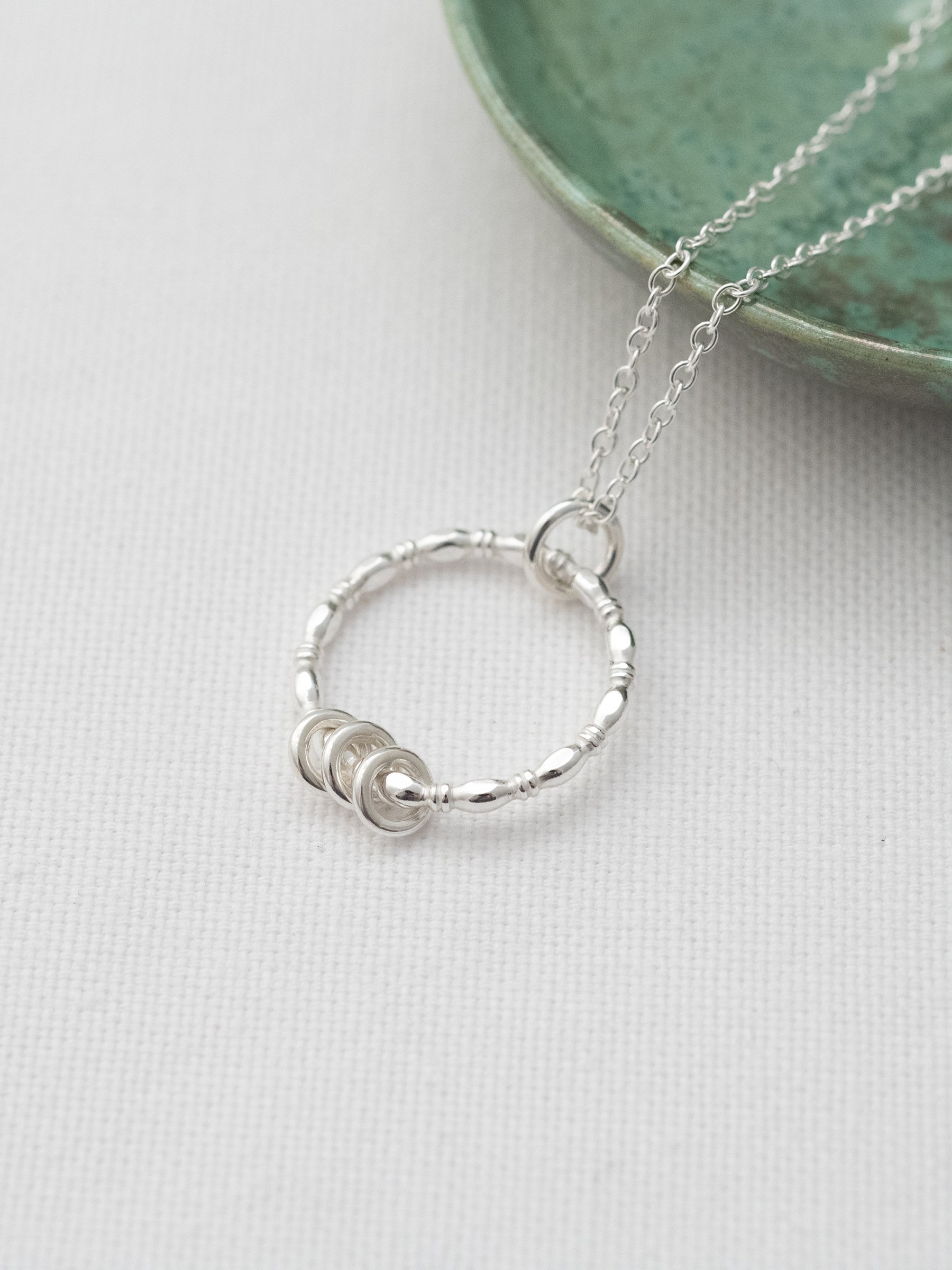 A silver three ring necklace displayed on white material with a handmade green jewellery dish in the background.