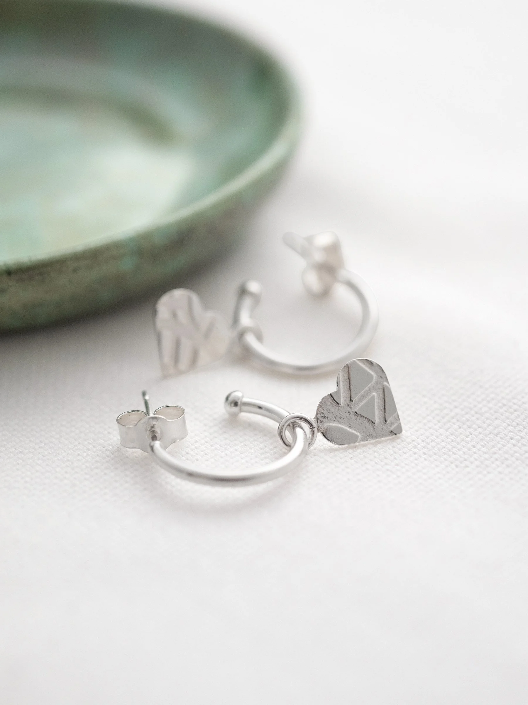 Sterling silver heart charm hoop stud earrings displayed on white material with a green jewellery dish in the background.