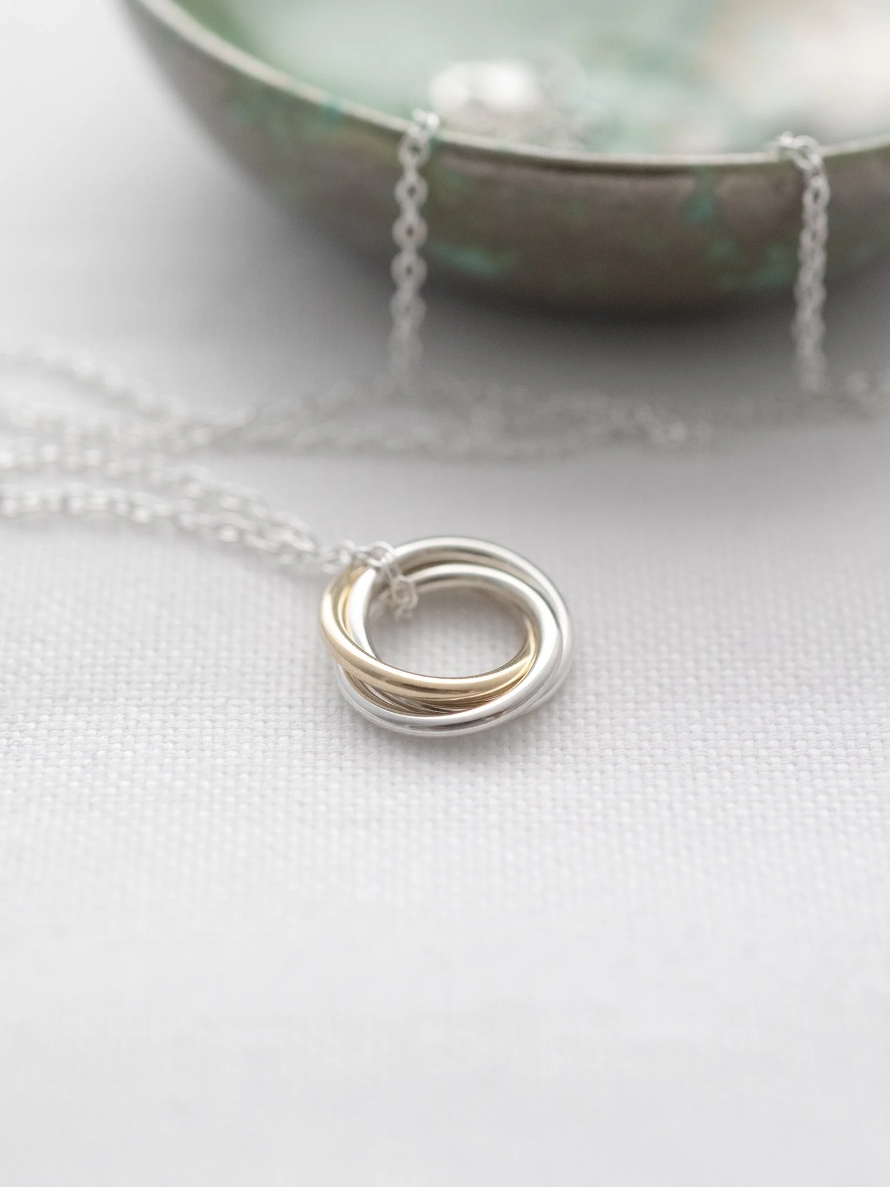 A close up of a gold and silver 3 ring necklace displayed on white material with a green jewellery dish in the background.