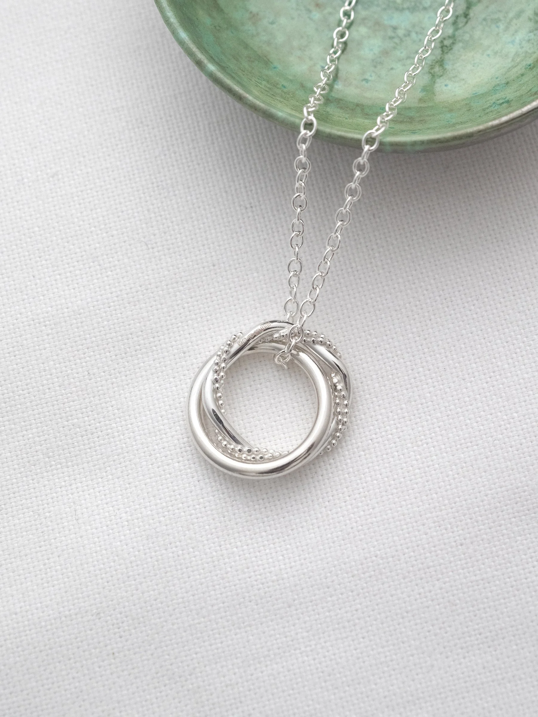 A hallmarked sterling silver two ring necklace for a 21st birthday displayed on white material with a green jewellery dish in the background.