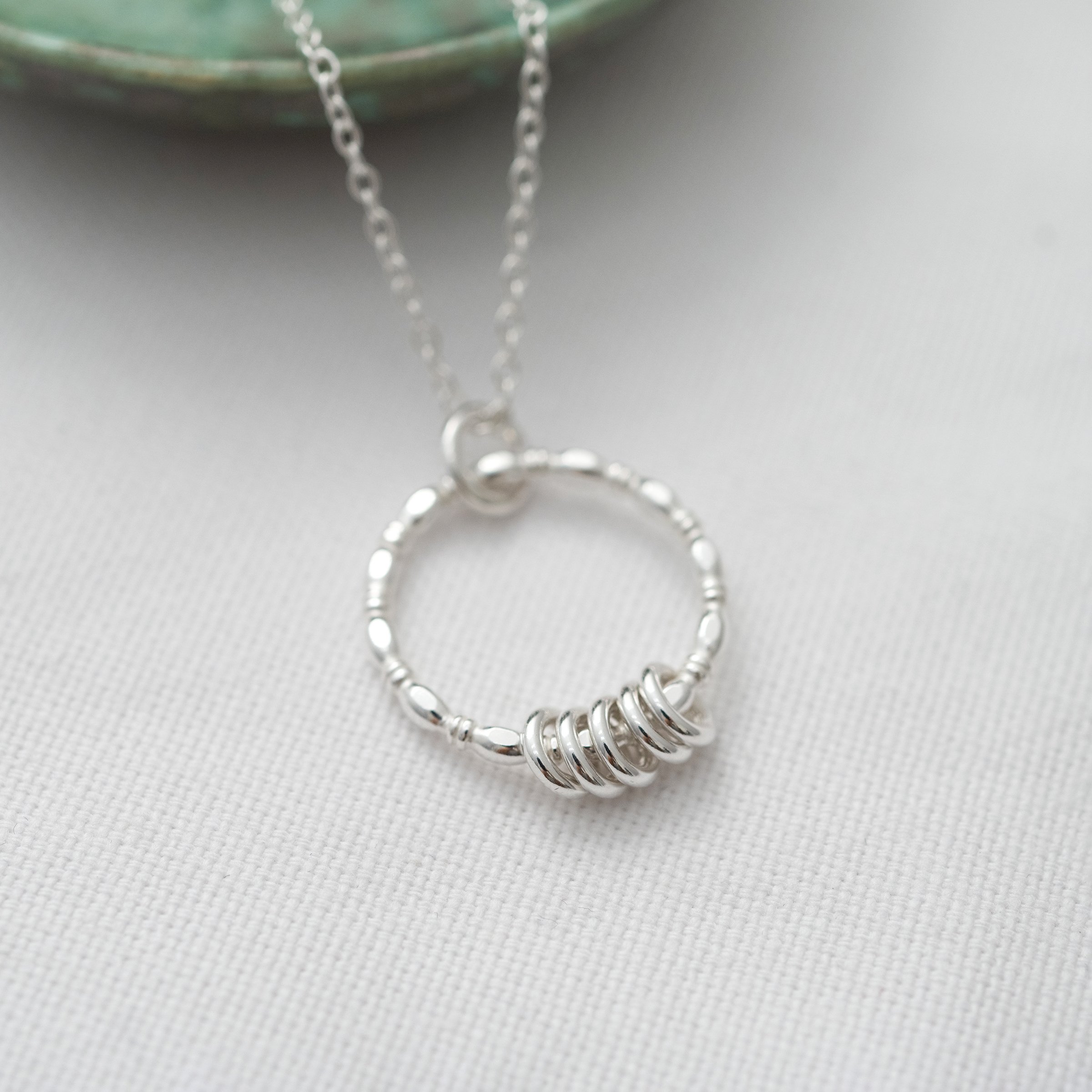 A silver 5 ring necklace displayed over white material with a green jewellery dish in the background.